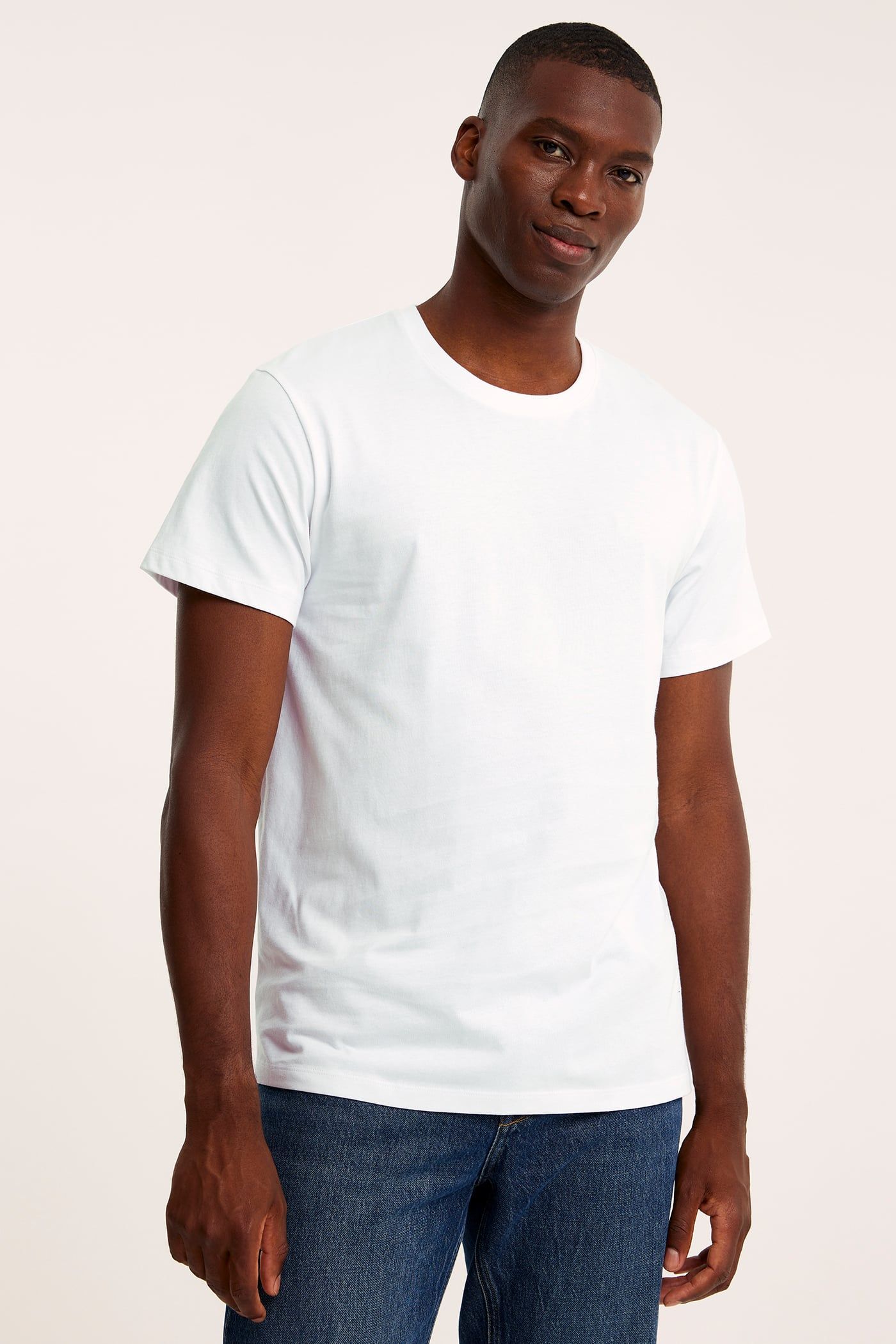 Framifrån: en man i en vit, kortärmad t-shirt med rund hals i bomull och blå jeans.
