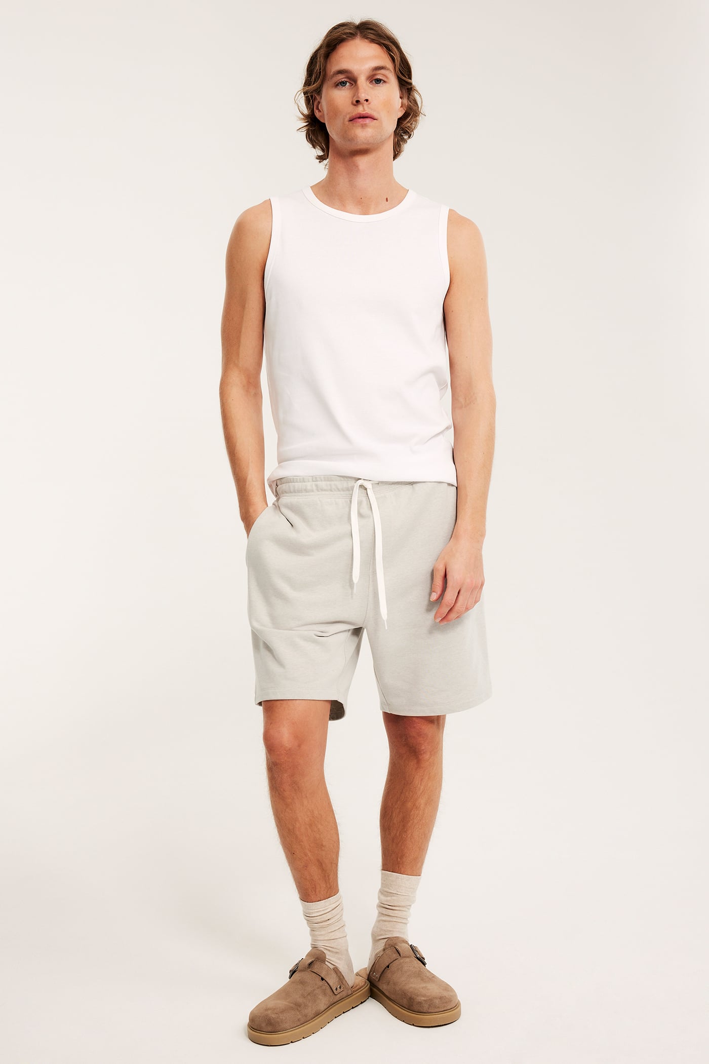 Framifrån, en man i melerade beiga mjukisshorts med dragsko, vitt linne och bruna tofflor.