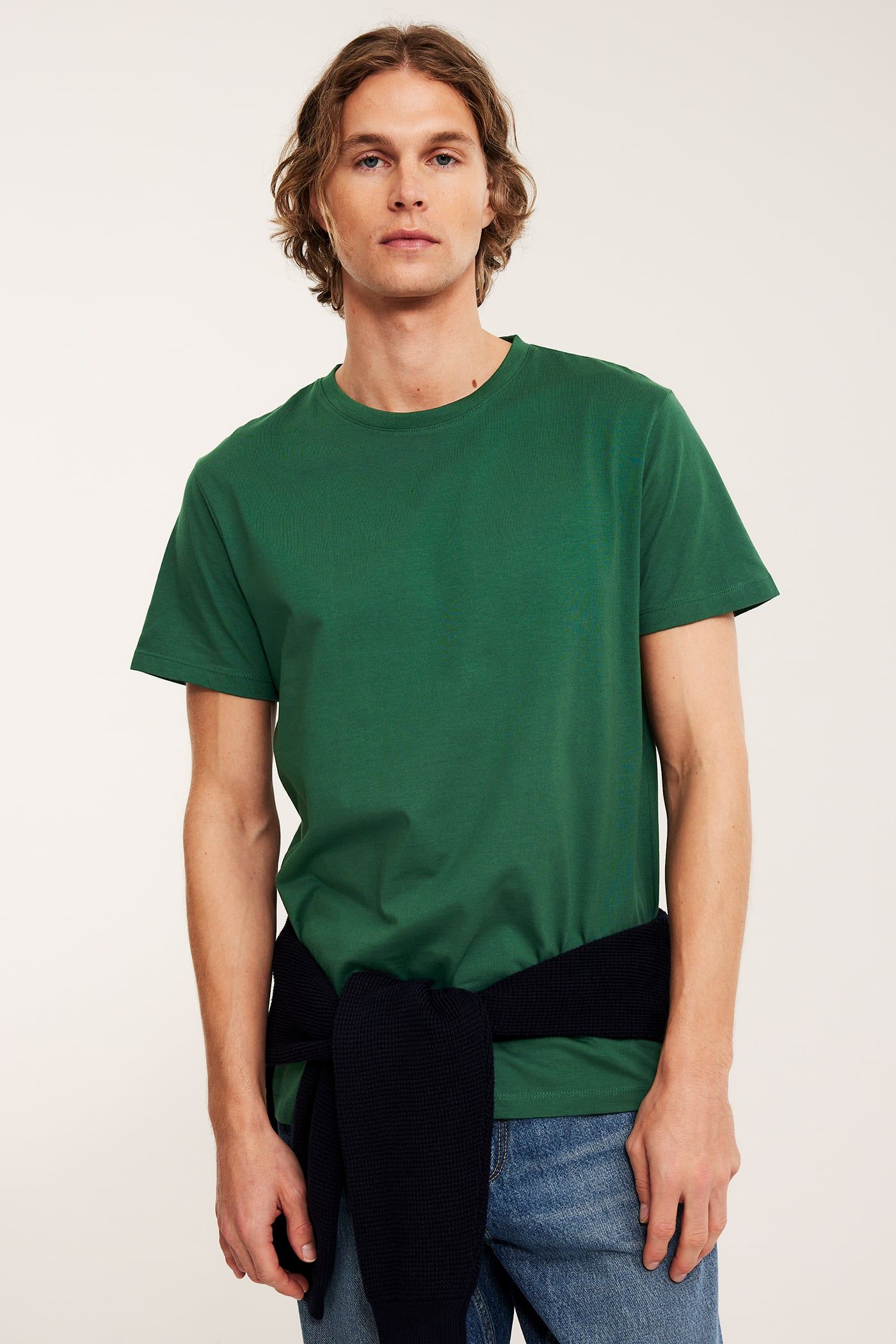Framifrån, man i mörkgrön bomulls-t-shirt, blåa jeans och mörkblå tröja knuten runt midjan.