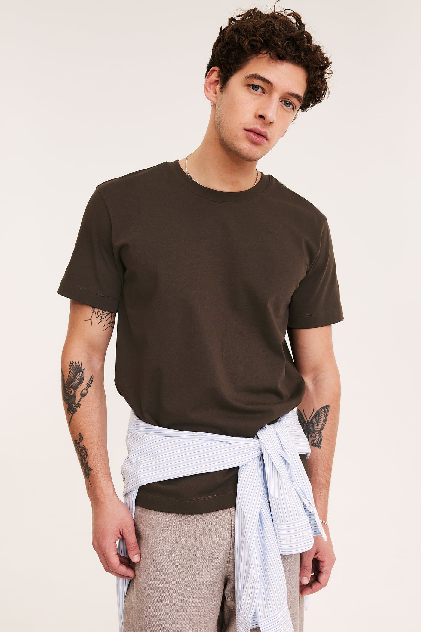 Framsida: Man i brun lös bomulls-O-hals t-shirt, beige shorts, och randig skjorta knuten i midjan.