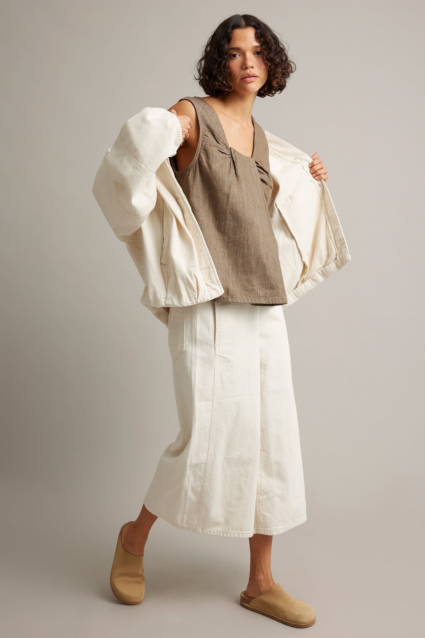 Kvinna bär brun ärmlös topp, krämvita vida byxor, krämvit öppen jacka, beige tofflor.