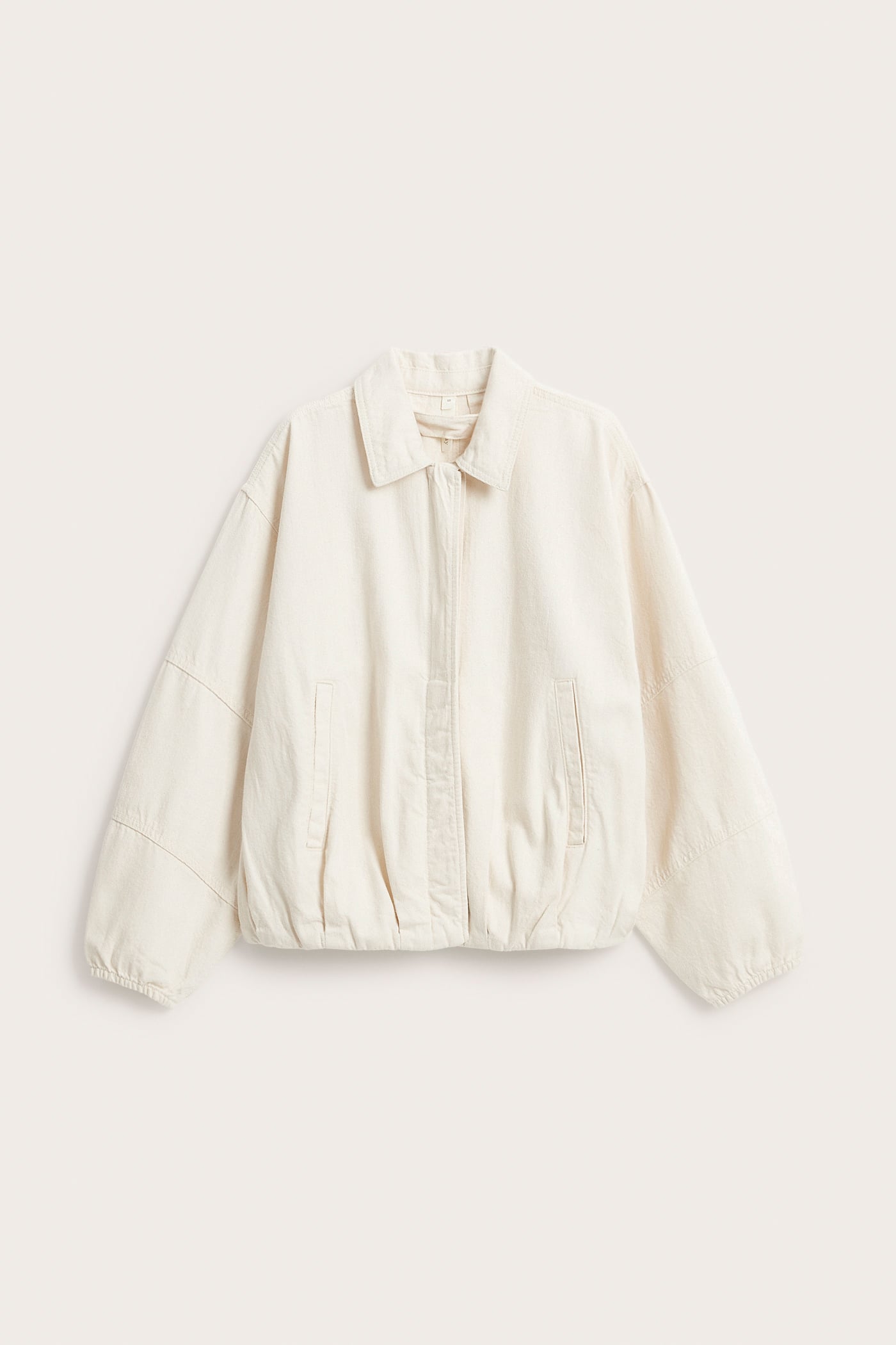 Framifrån: Ljusbeige bomberjacka i bomullsdenim med krage, dolda knappar och stolpfickor fram.