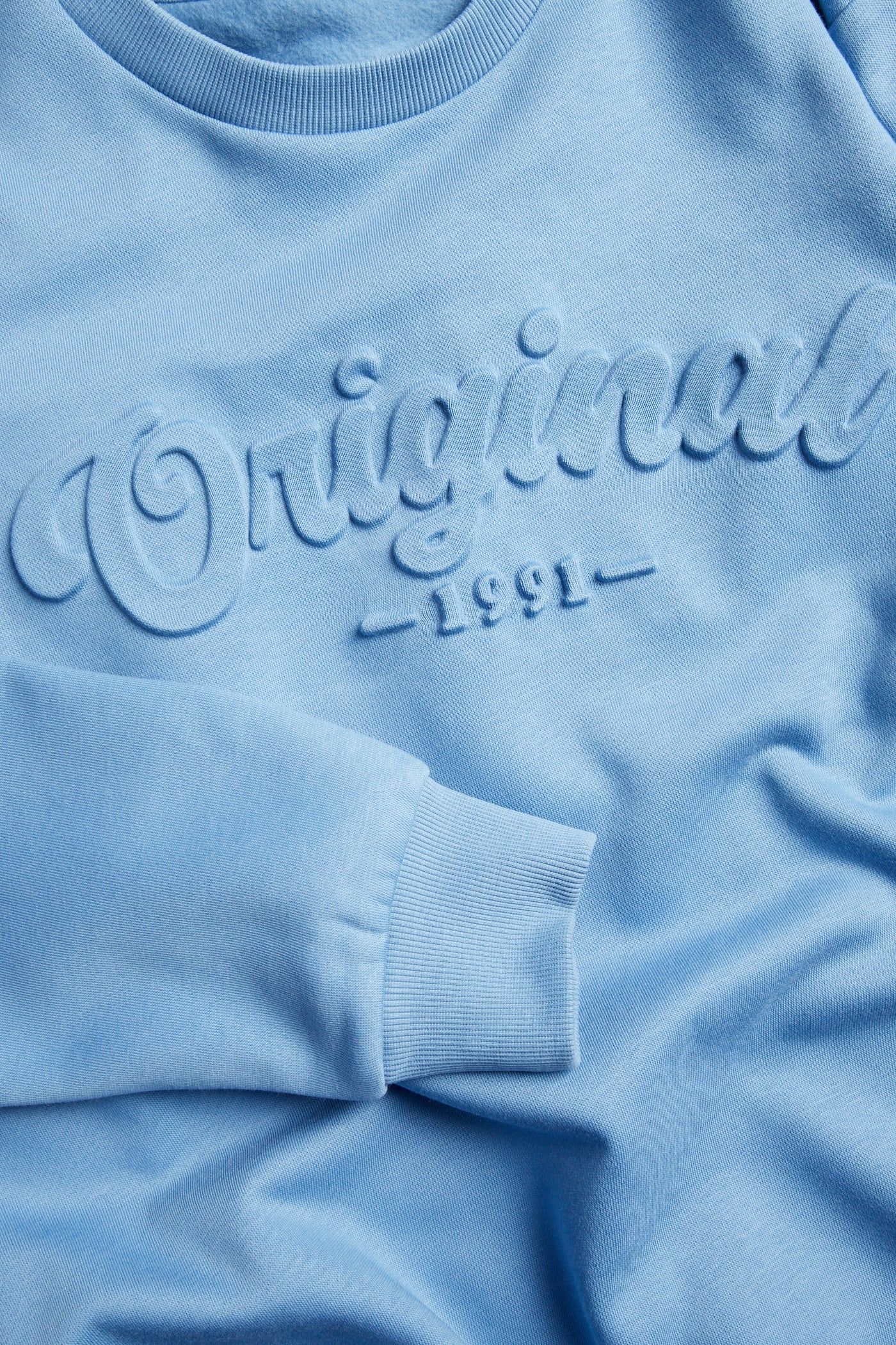 Zbliżenie na jasnoniebieską bluzę z wytłoczonym napisem „Originals 1991” z przodu.