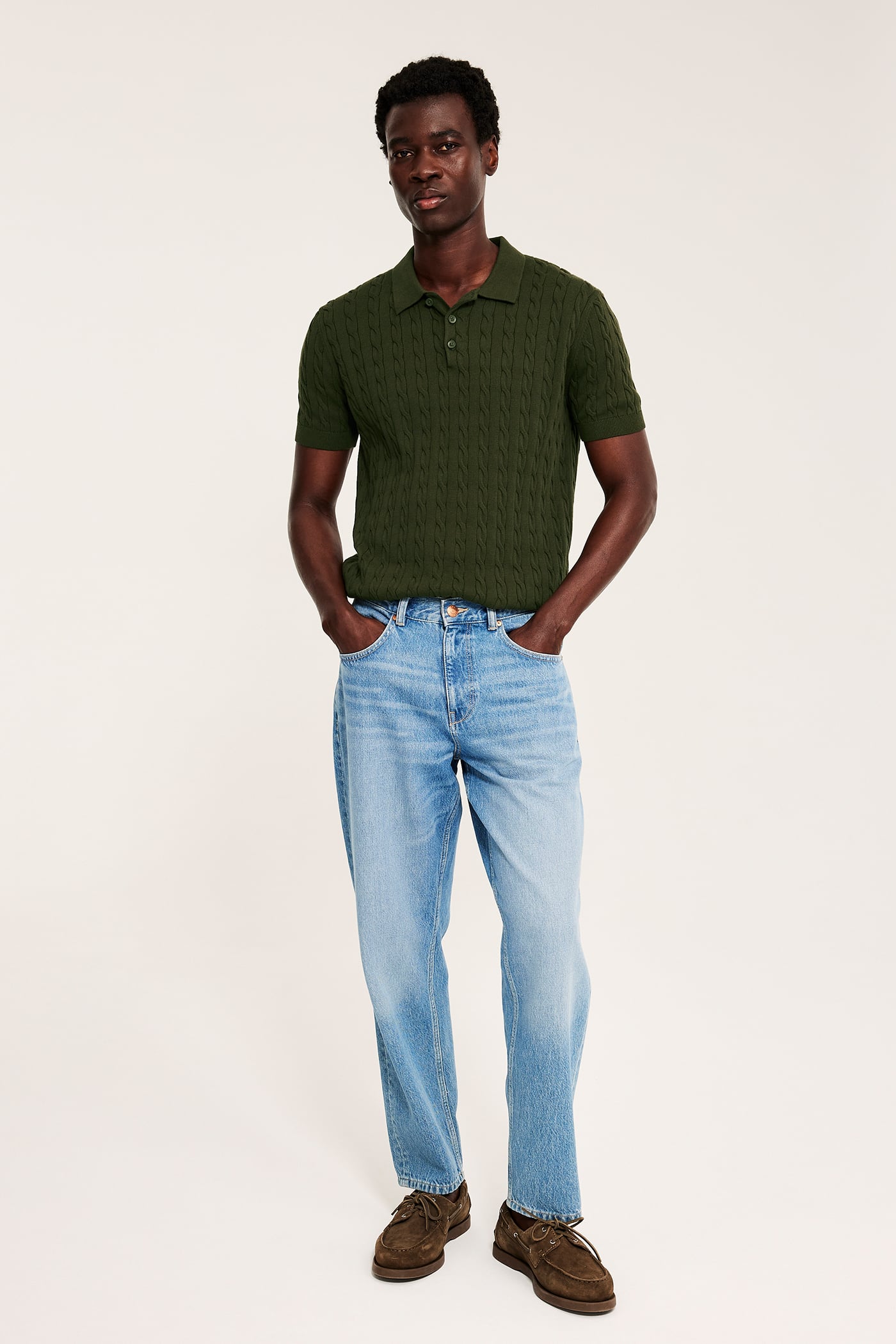 Framifrån, manlig modell i mörkgrön kabelstickad pikétröja, ljusblå jeans och bruna seglarskor.