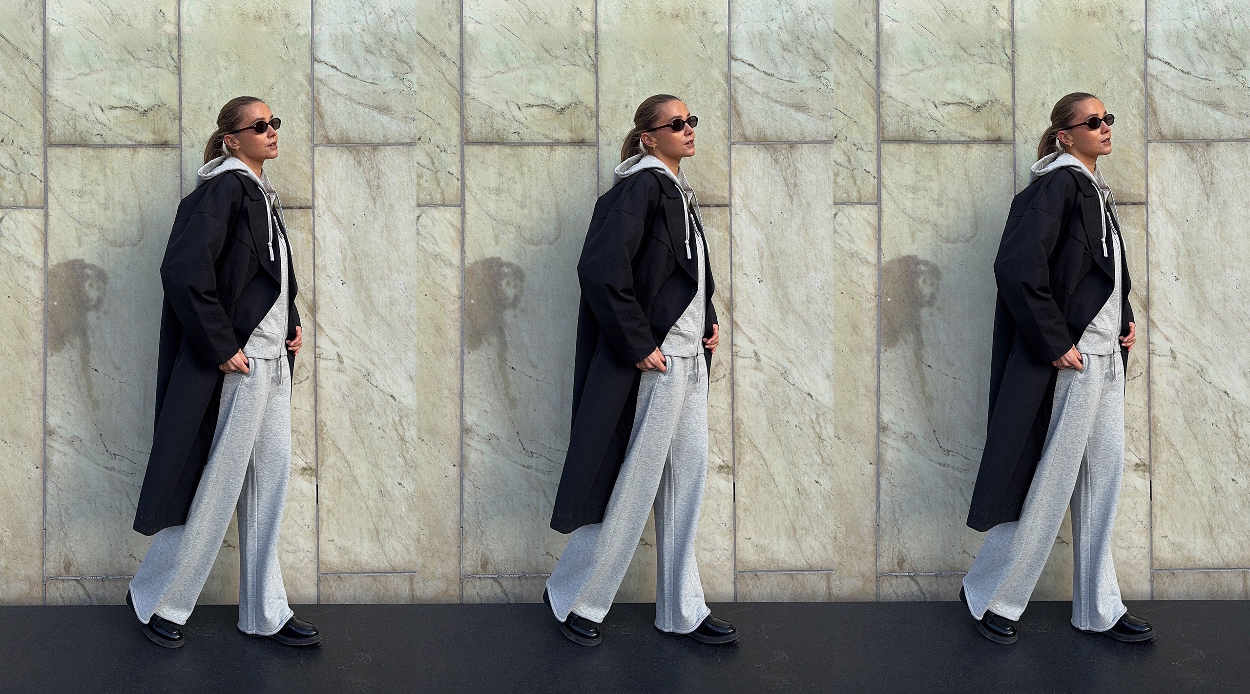 Kobieta w szarym zestawie loungewear i czarnym płaszczu na tle kamiennej ściany.