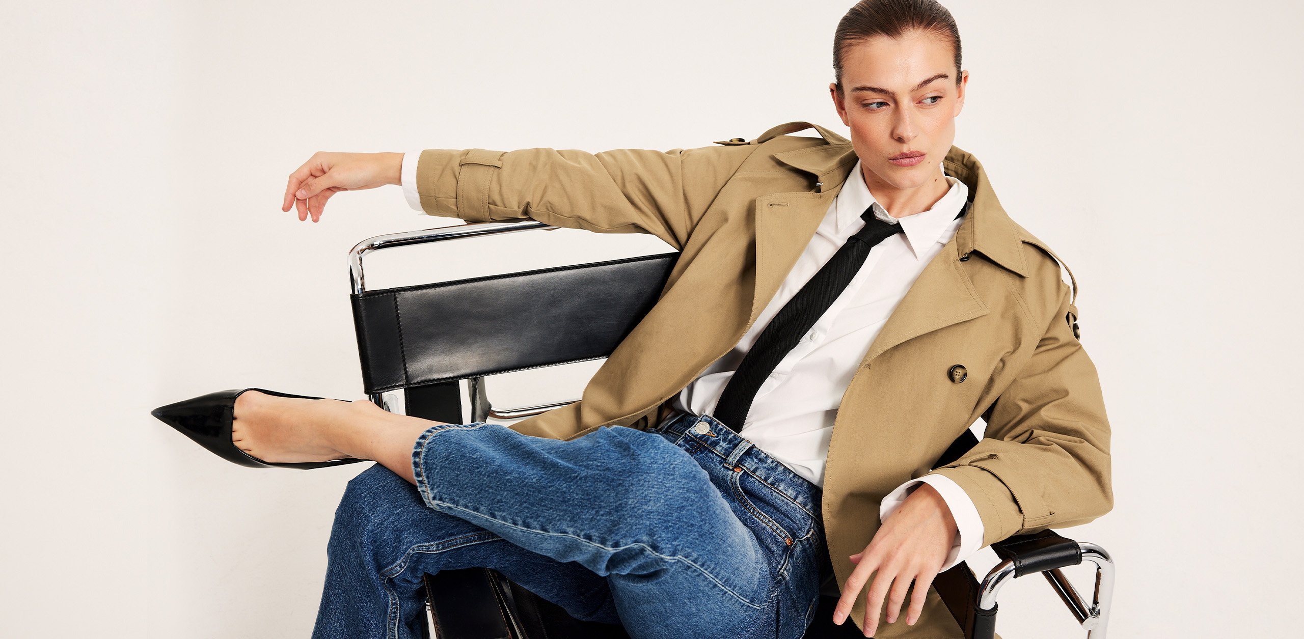 En kvinne kledd i en kort trenchcoat, jeans, skjorte og slips.