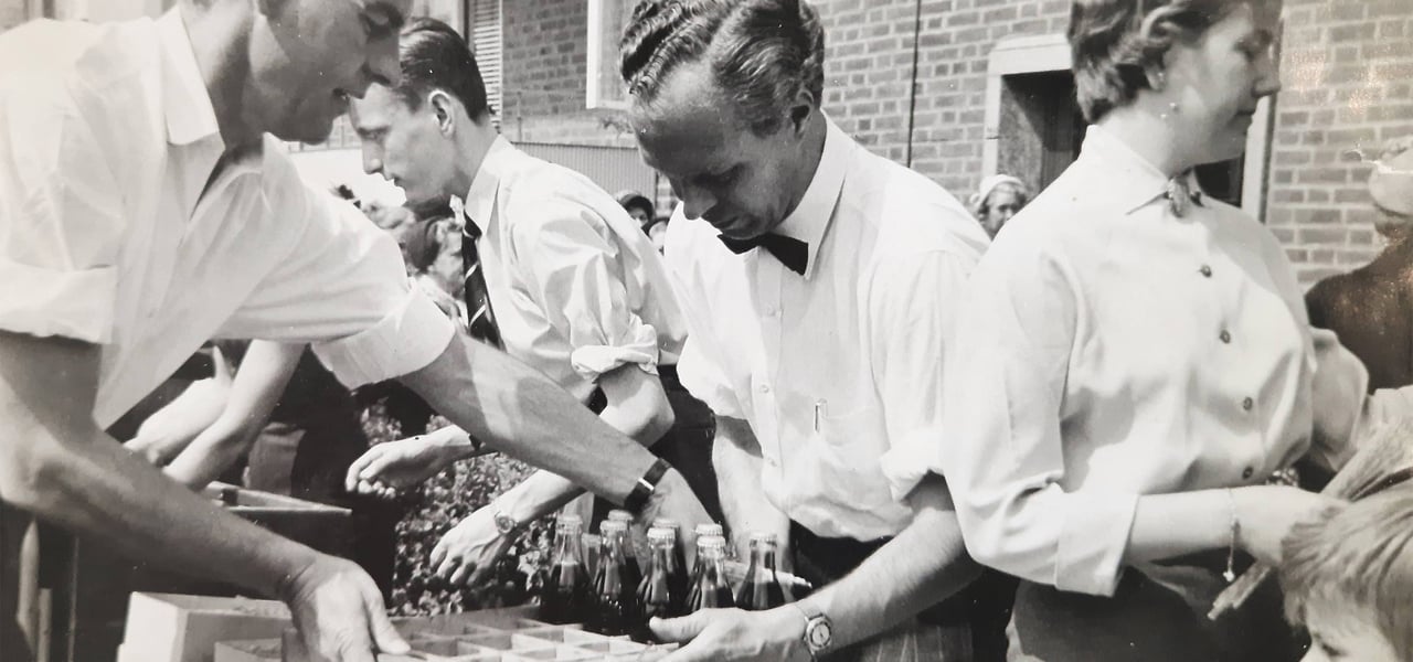 Czarno-białe zdjęcie, na którym mężczyźni w białych koszulach podają Coca-Colę.