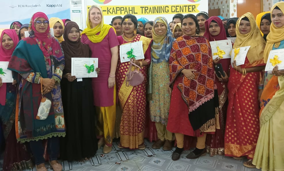Hymyilevät naiset värikkäissä juhla-asuissa pitelevät diplomeja kyltin edessä, jossa lukee "Kappahl Training Center".