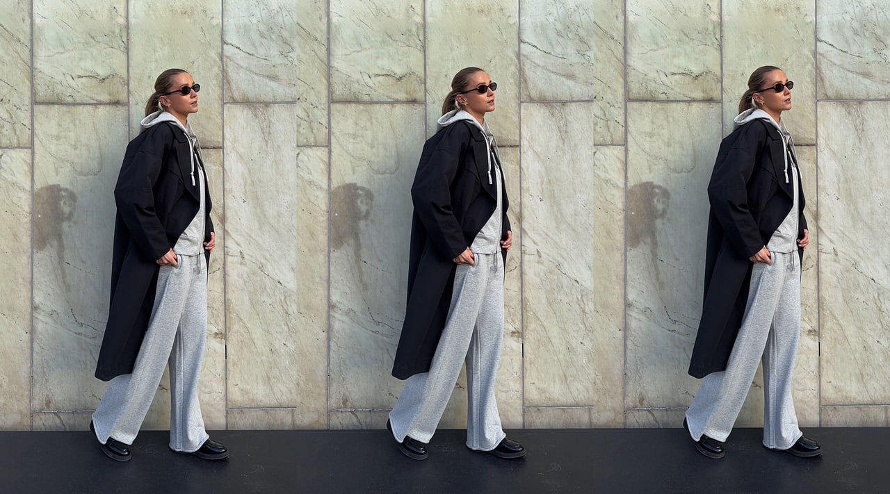 Kobieta w szarym zestawie loungewear i czarnym płaszczu na tle kamiennej ściany. 