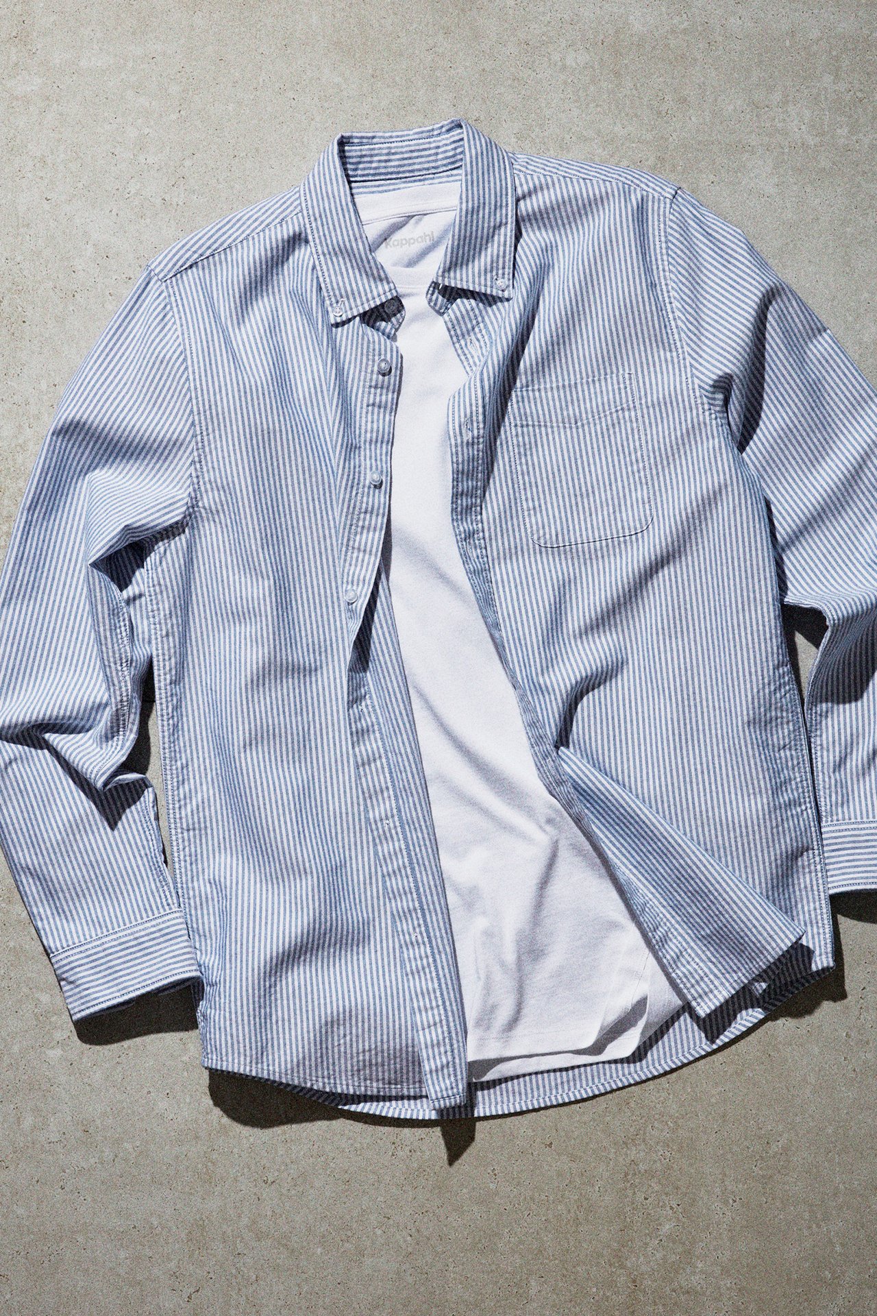 Niebiesko-biała koszula w paski z białą koszulką pod spodem. Zdjęcie wykonane na tle betonowej podłogi. 