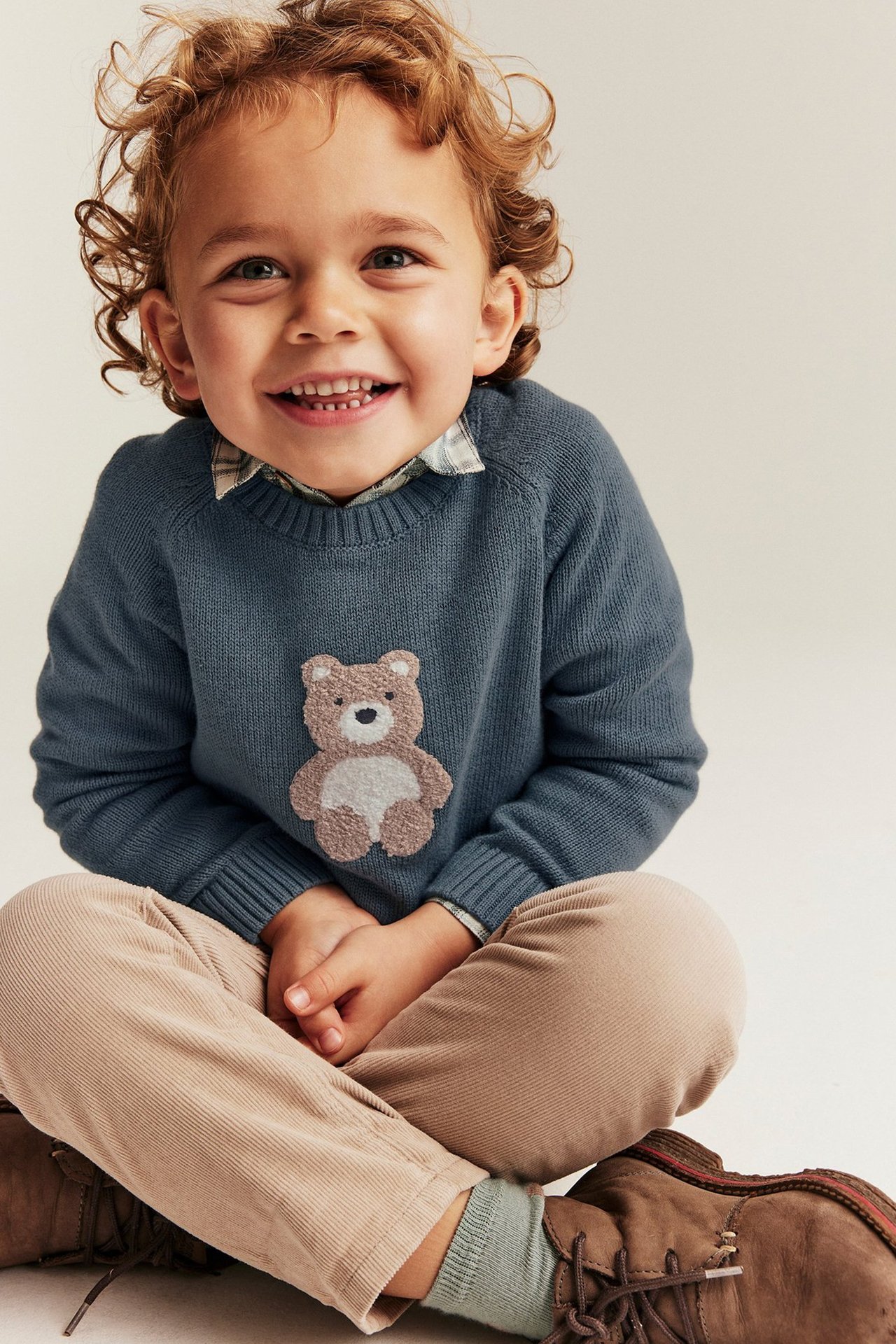 Małe dziecko siedzące na podłodze z szerokim uśmiechem, ubrane w sweter i spodnie od Newbie.