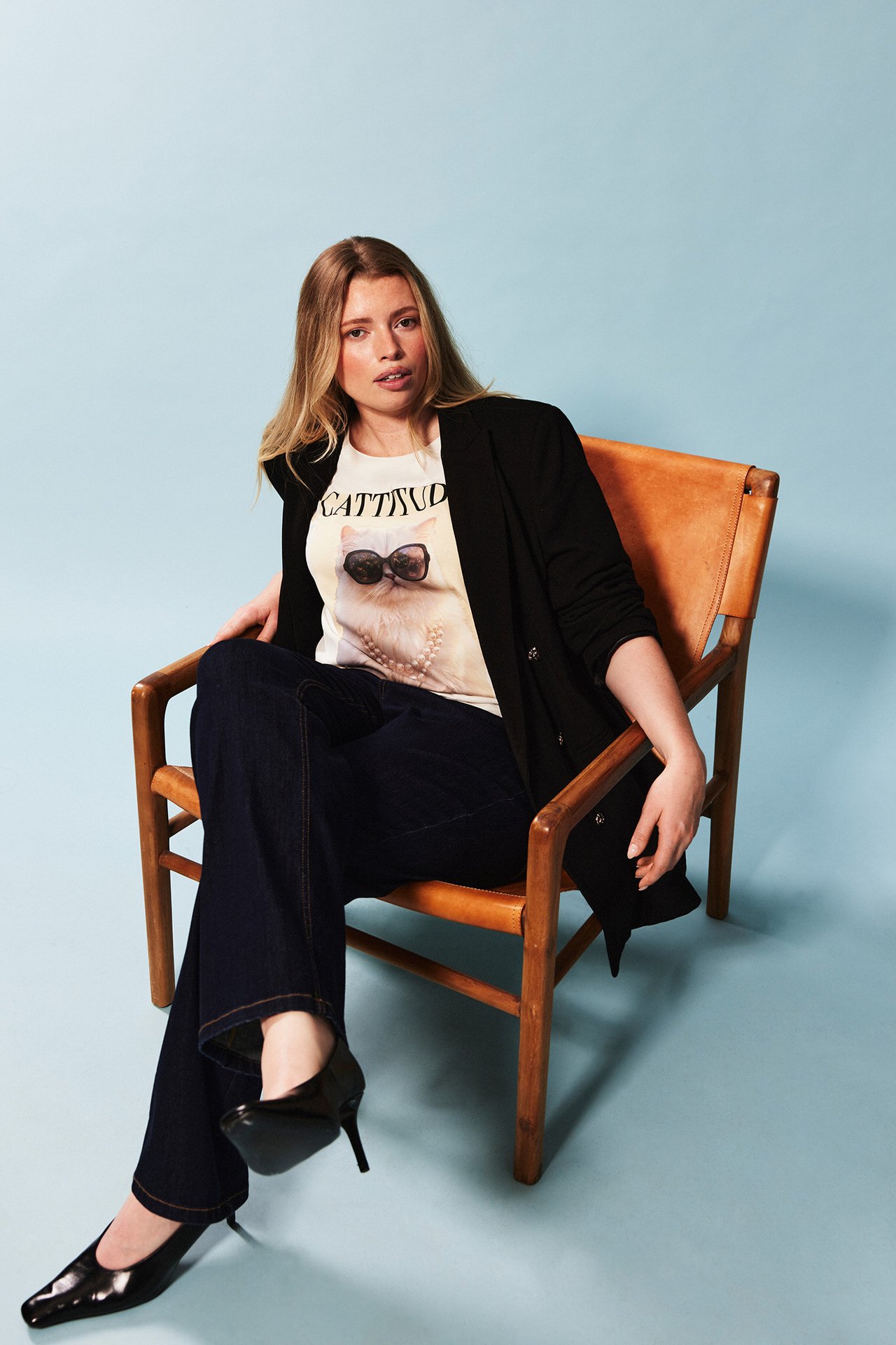 Person i svart kavaj, grafisk t-shirt med kattmotiv, mörkblå jeans och svarta pumps sitter i en brun läderfåtölj.