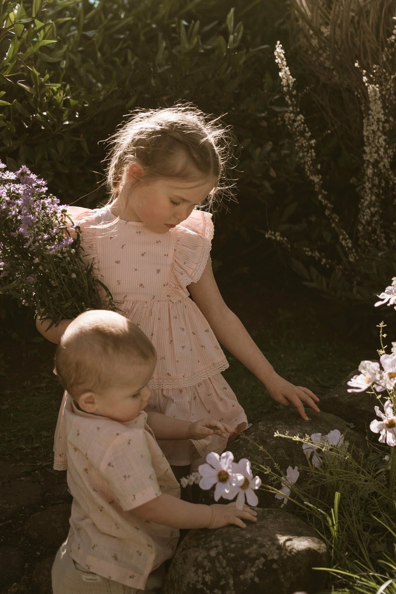 Children wearing light summer clothing picking flowers outdoors in warm evening light.