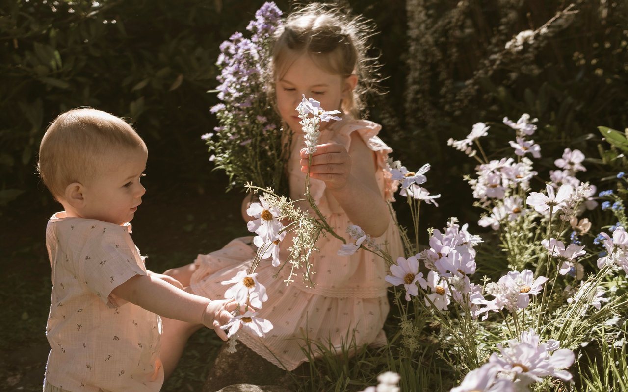 Two children in soft pink summer outfits sitting among wildflowers in a sunlit garden.