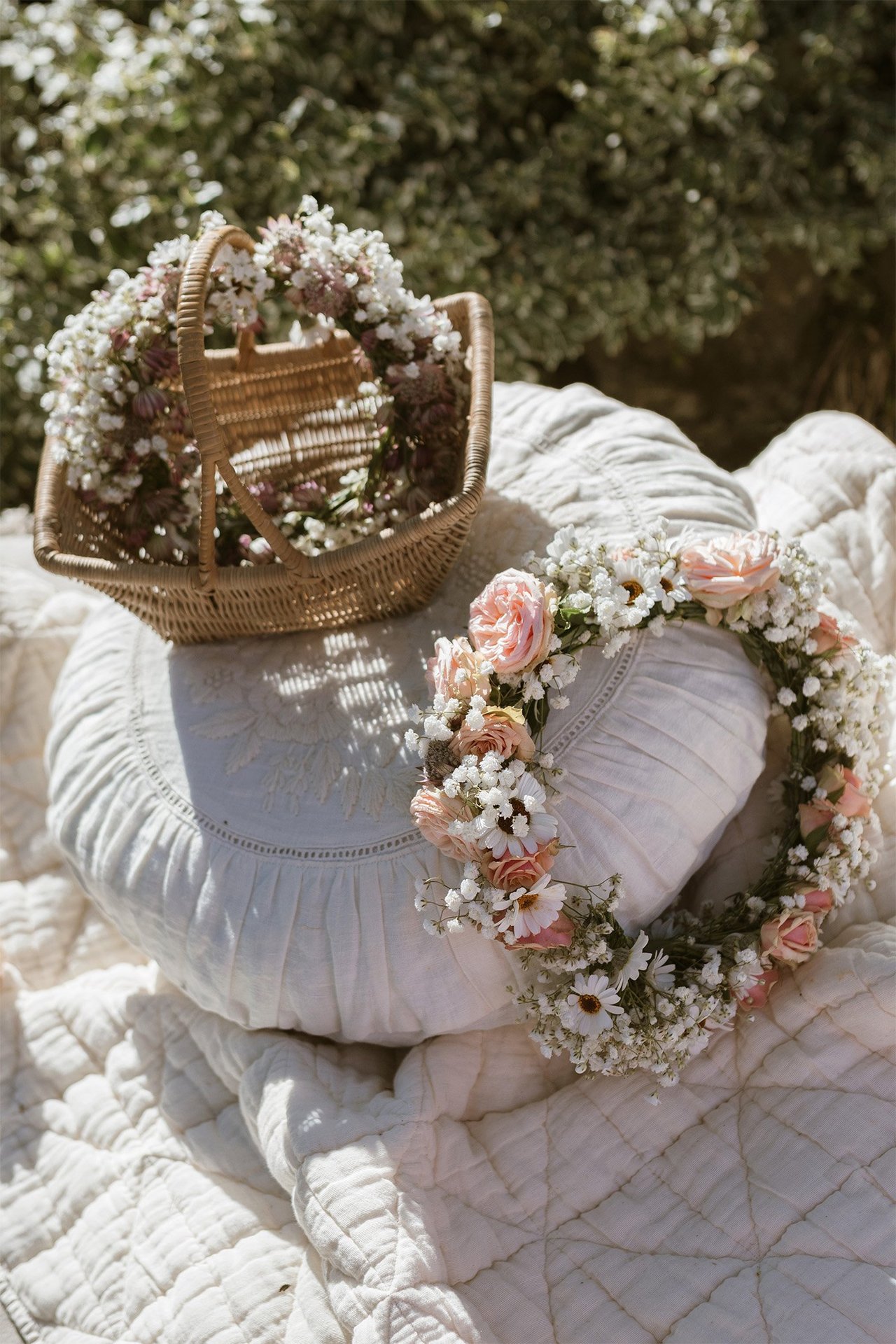 A floral wreath and woven basket resting on soft white fabric in a sunlit garden.