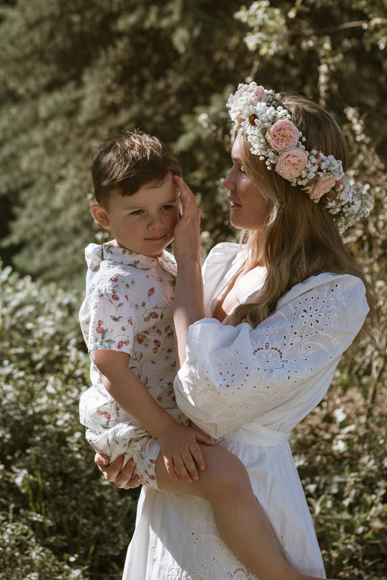 An adult wearing a white dress and flower crown holding a child outdoors in summer greenery.