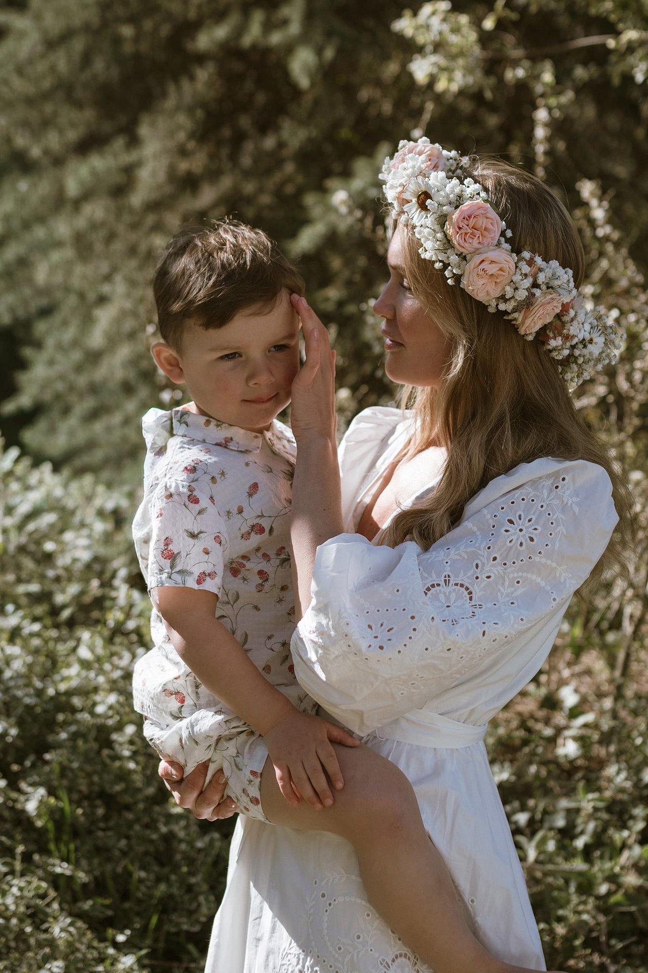 An adult wearing a white dress and flower crown holding a child outdoors in summer greenery.