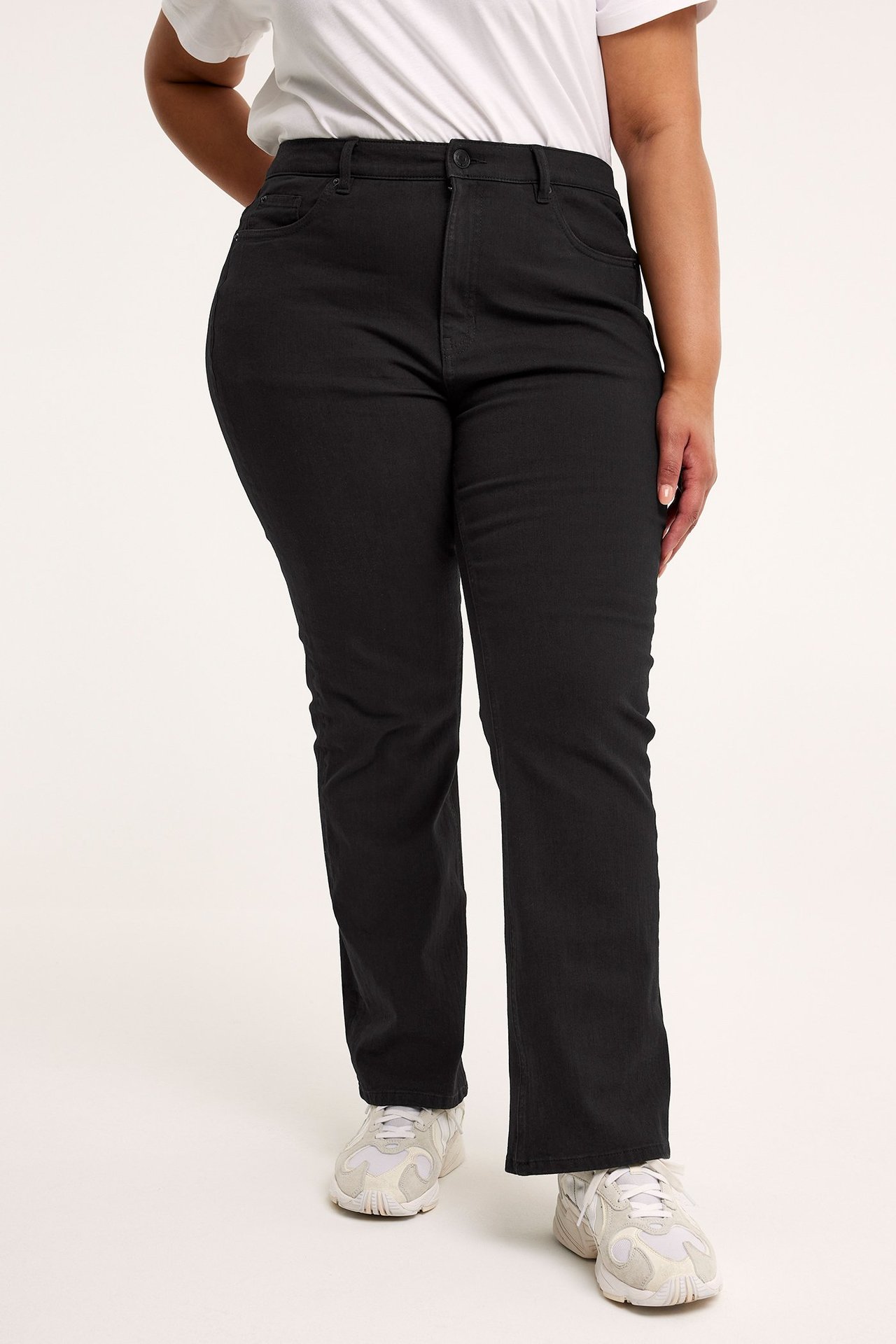 Frontvy av en kvinna i svarta bootcut jeans med hög midja, vit t-shirt och vita sneakers.