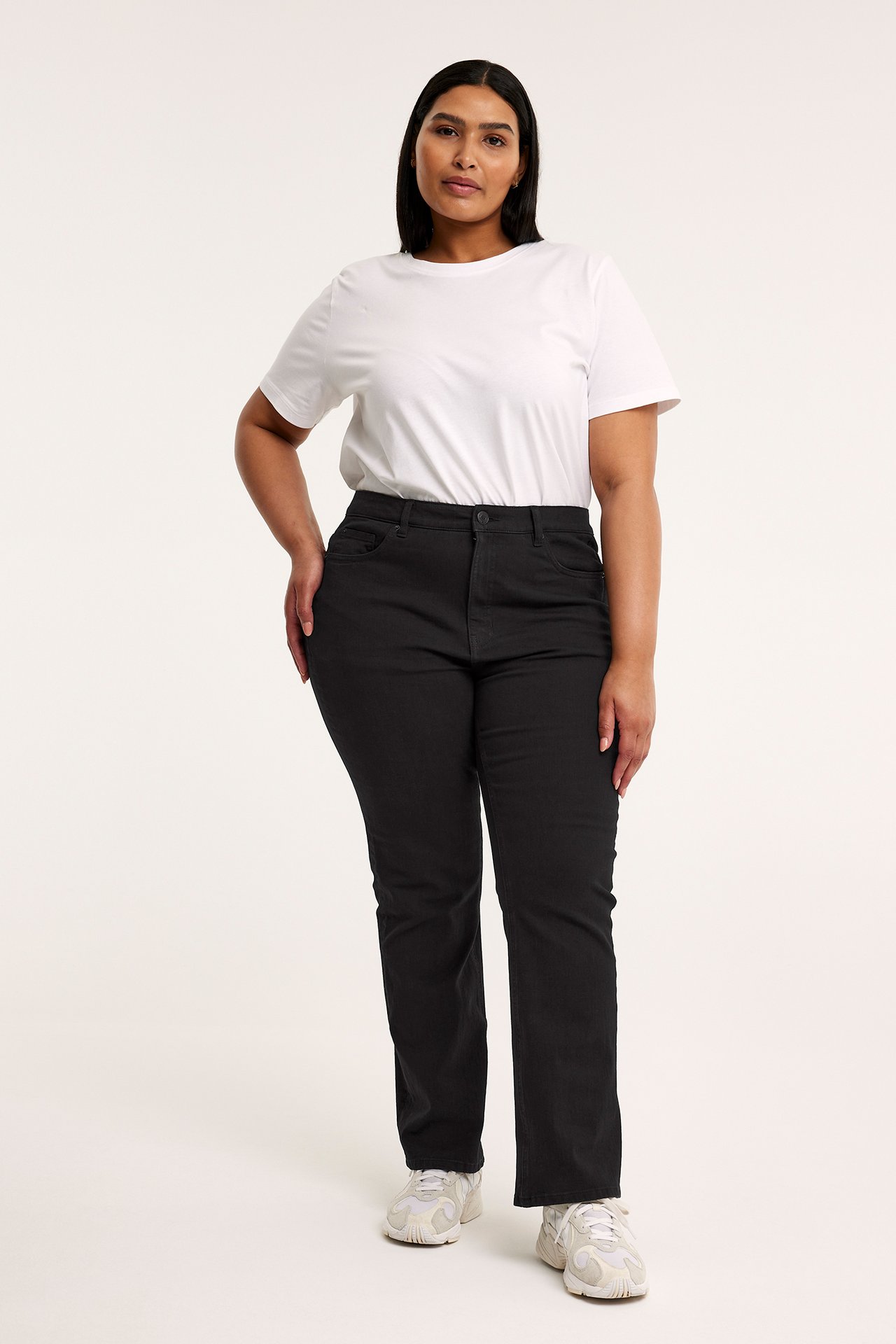 Framifrån, kvinna i svarta bootcut-jeans med hög midja, vit t-shirt och vita sneakers.