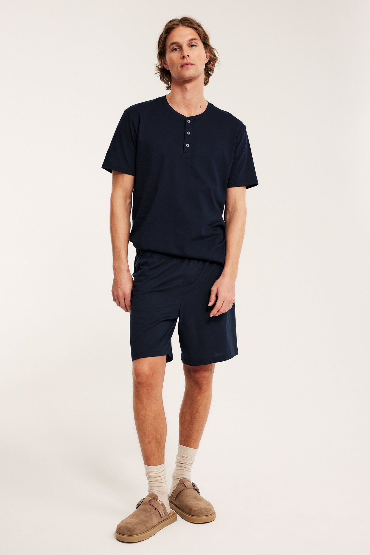 Framifrån, en man i mörkblå jersey pyjamasshorts, matchande Henley-t-shirt och beige tofflor.