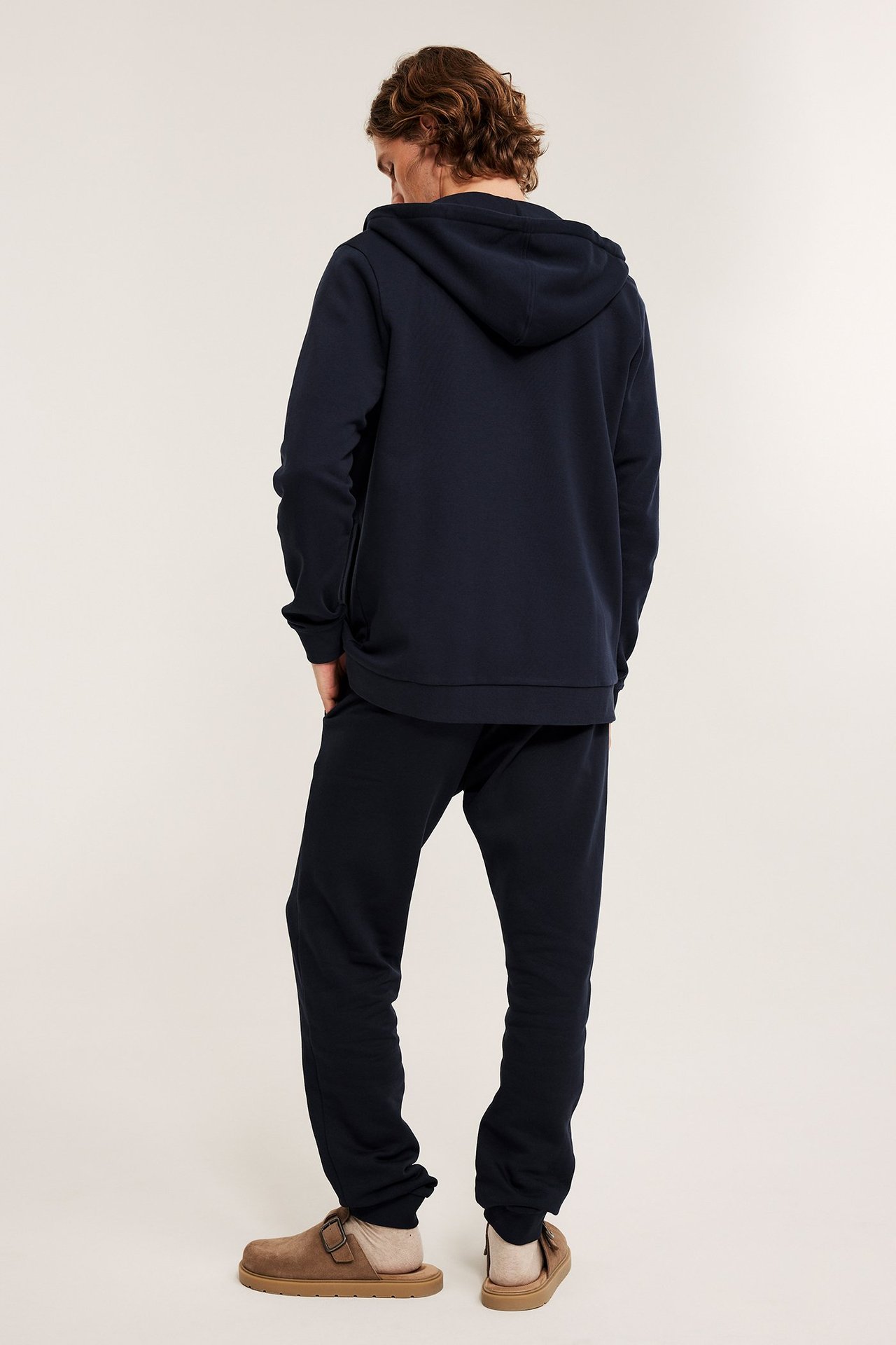 Takakuva: Mies tummansinisissä collegehousuissa, yhteensopivassa hupparissa ja ruskeissa tossuissa.