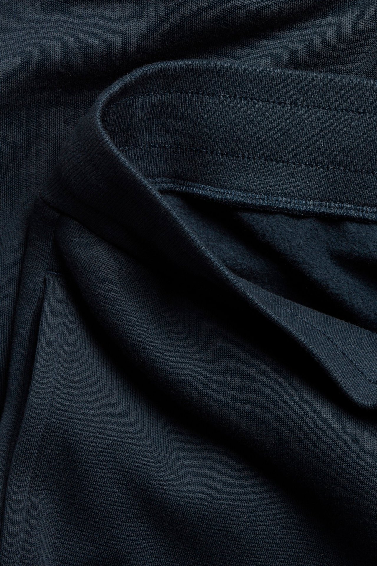 Lähikuva tummansinisistä collegehousuista, joissa näkyy leveä joustovyötärö, sivutasku ja harjattu sisäpuoli.