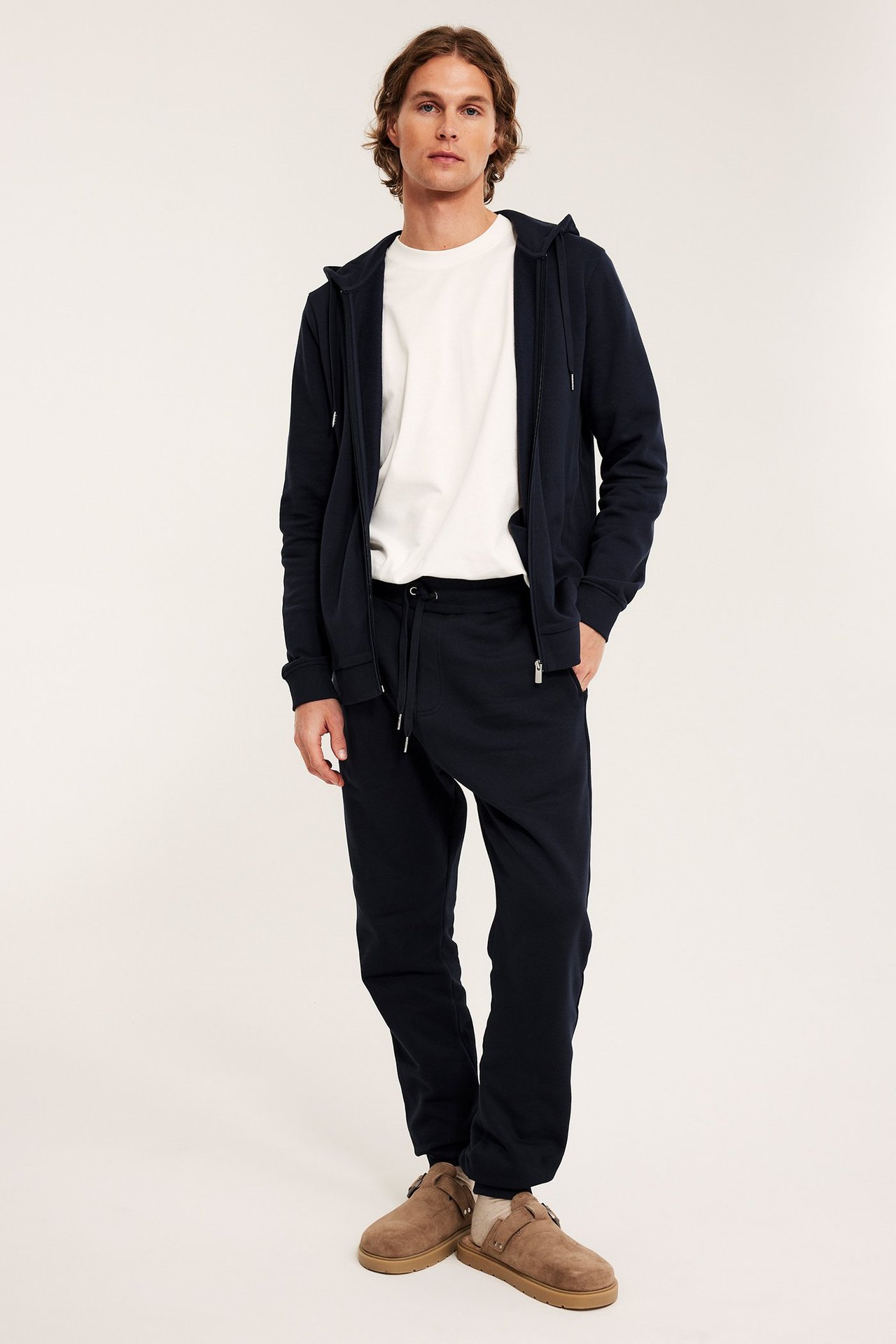 Etunäkymä, mies yllään tummansiniset collegehousut, yhteensopiva vetoketjuhuppari, valkoinen t-paita ja beiget tohvelit.