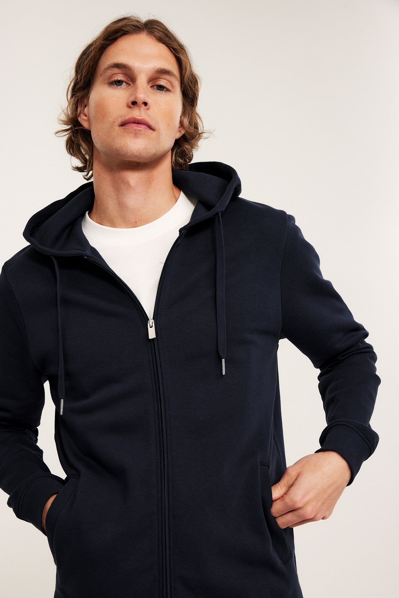 Framsiden av en mann i mørkeblå hettegenser med glidelås over hvit t-skjorte, med hendene i lommene.