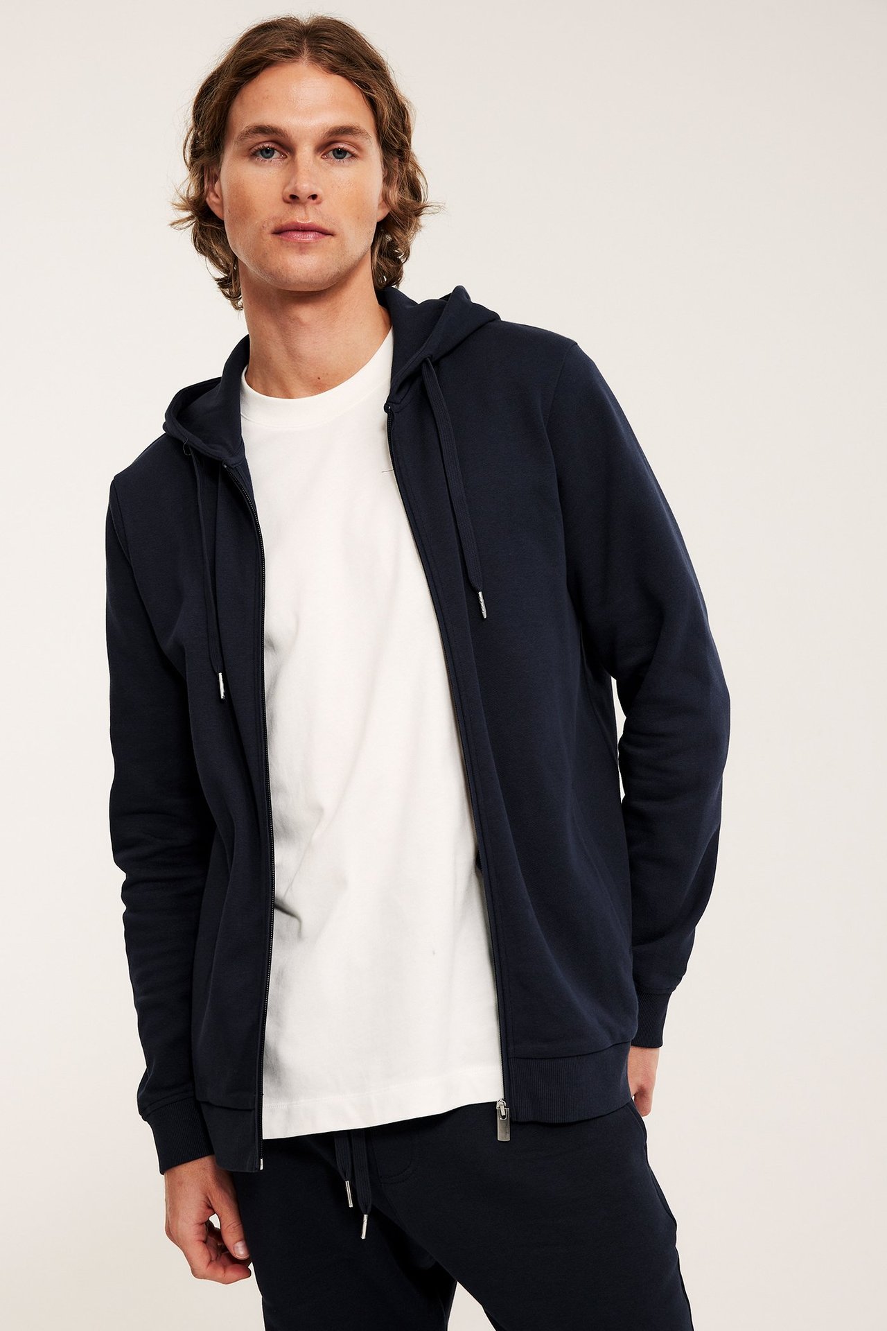 Edestä: mies avonaisessa tummansinisessä vetoketjuhupparissa valkoisen t-paidan ja tummansinisten collegehousujen kanssa.