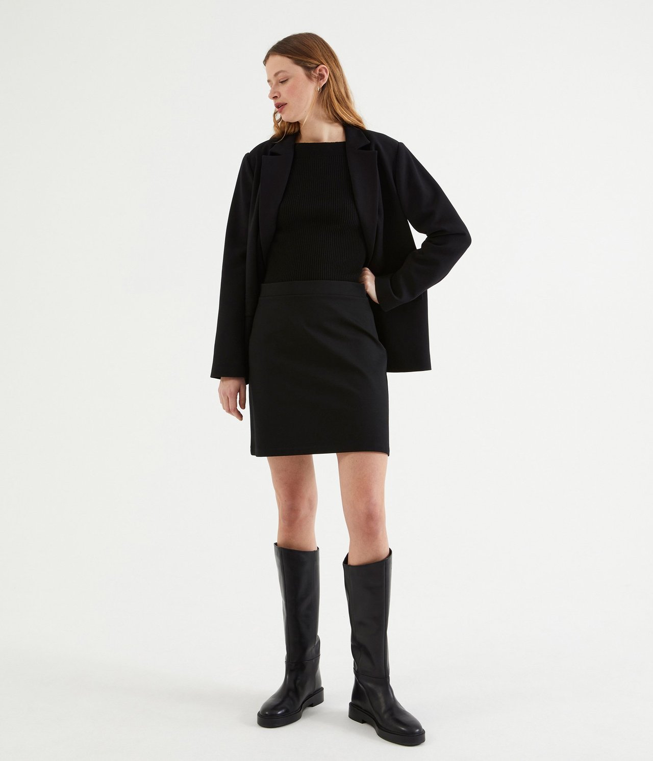 Widok z przodu kobiety w czarnej spódnicy ponte roma, prążkowanym topie, marynarce i kozakach.