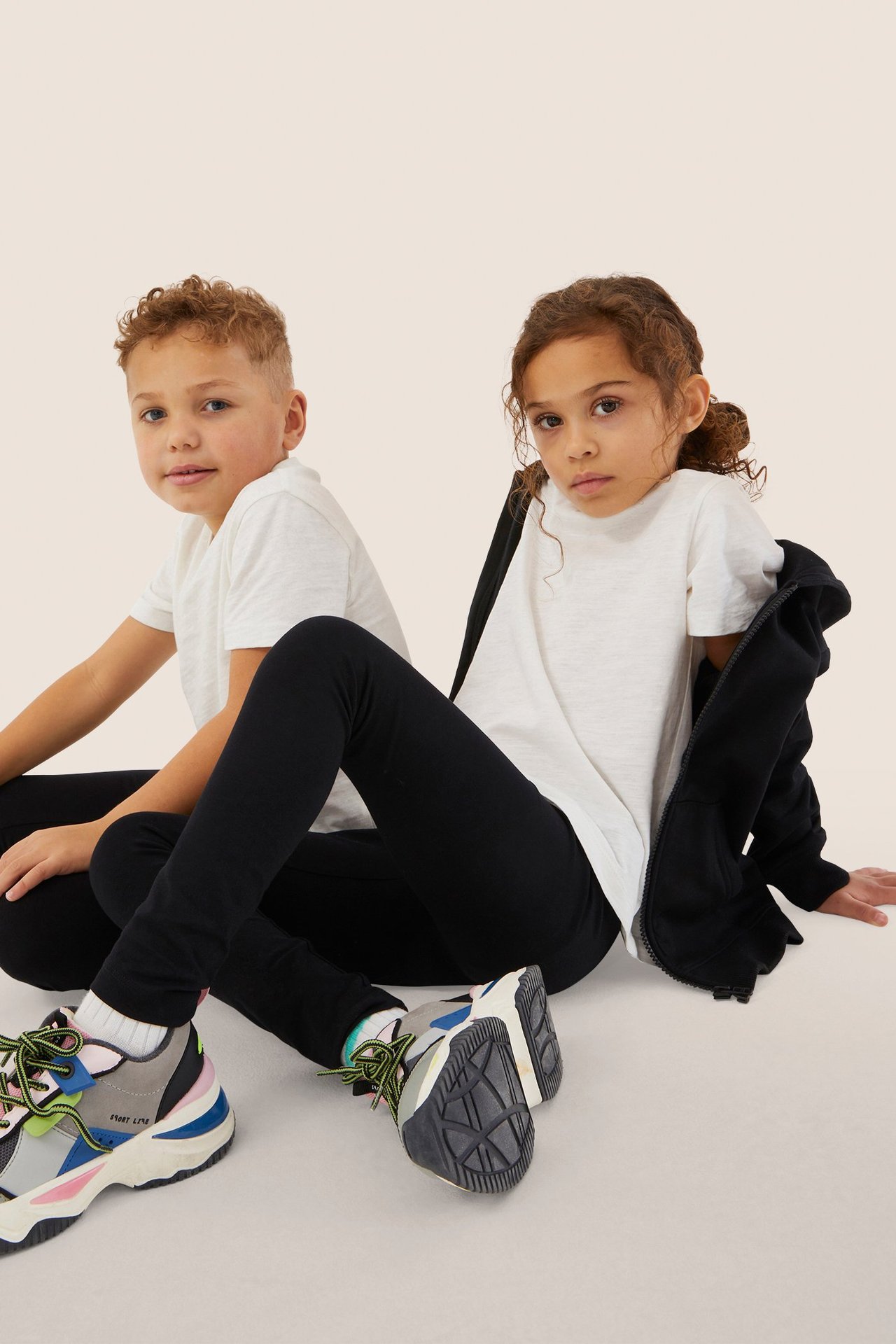Frontvisning: To barn i svarte leggings, hvite t-skjorter og fargerike joggesko. Jenta med svart hettegenser.