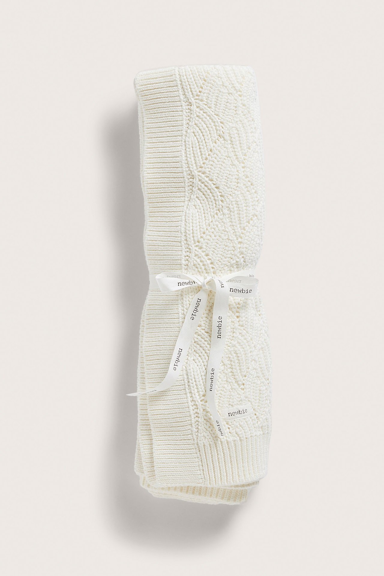 Framifrån: Offwhite stickad babyfilt med jacquardmönster, knuten med ett 'Newbie'-band.