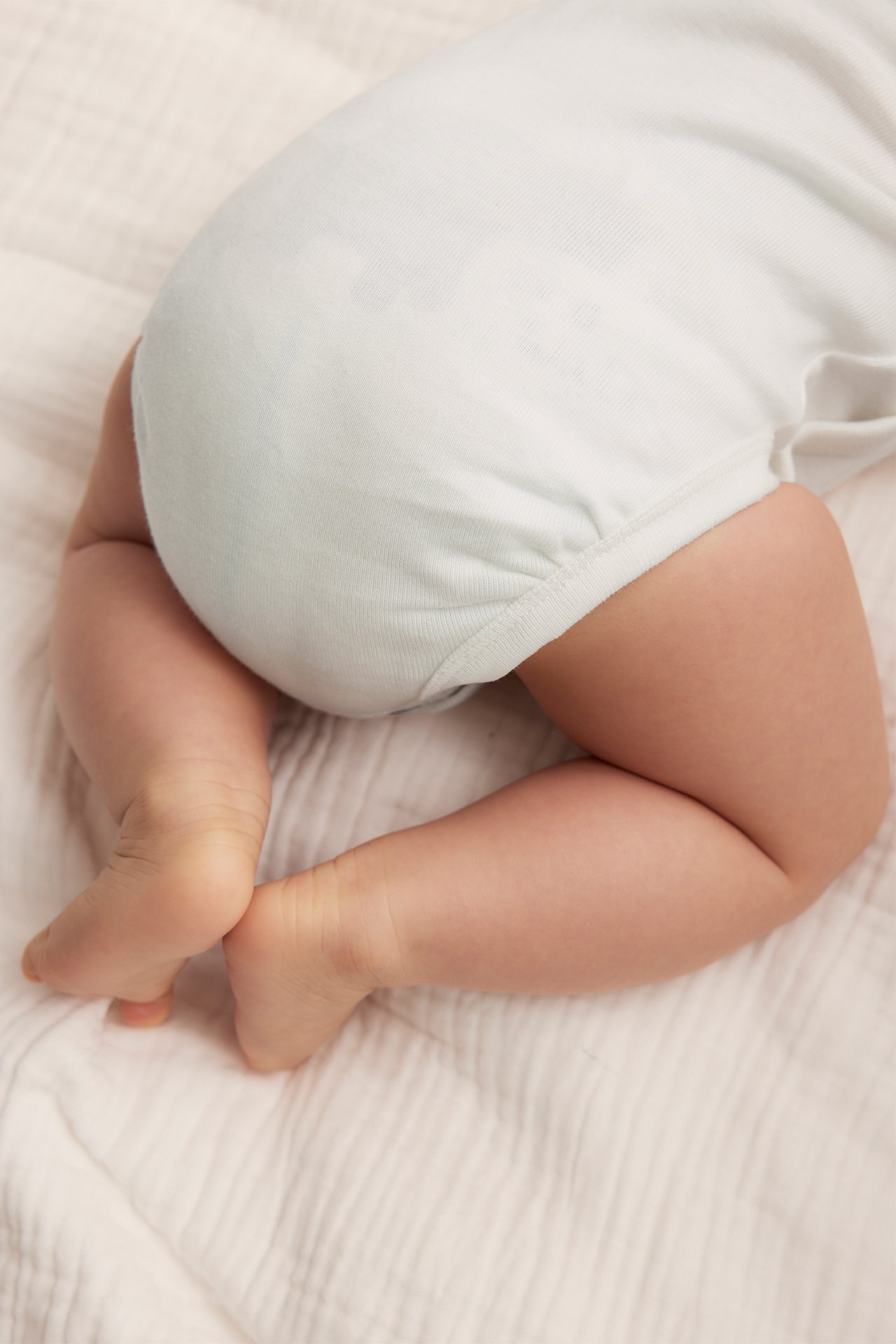 Bakfra, baby i en offwhite bomulls-omslagsbody, liggende på et lyst teppe.