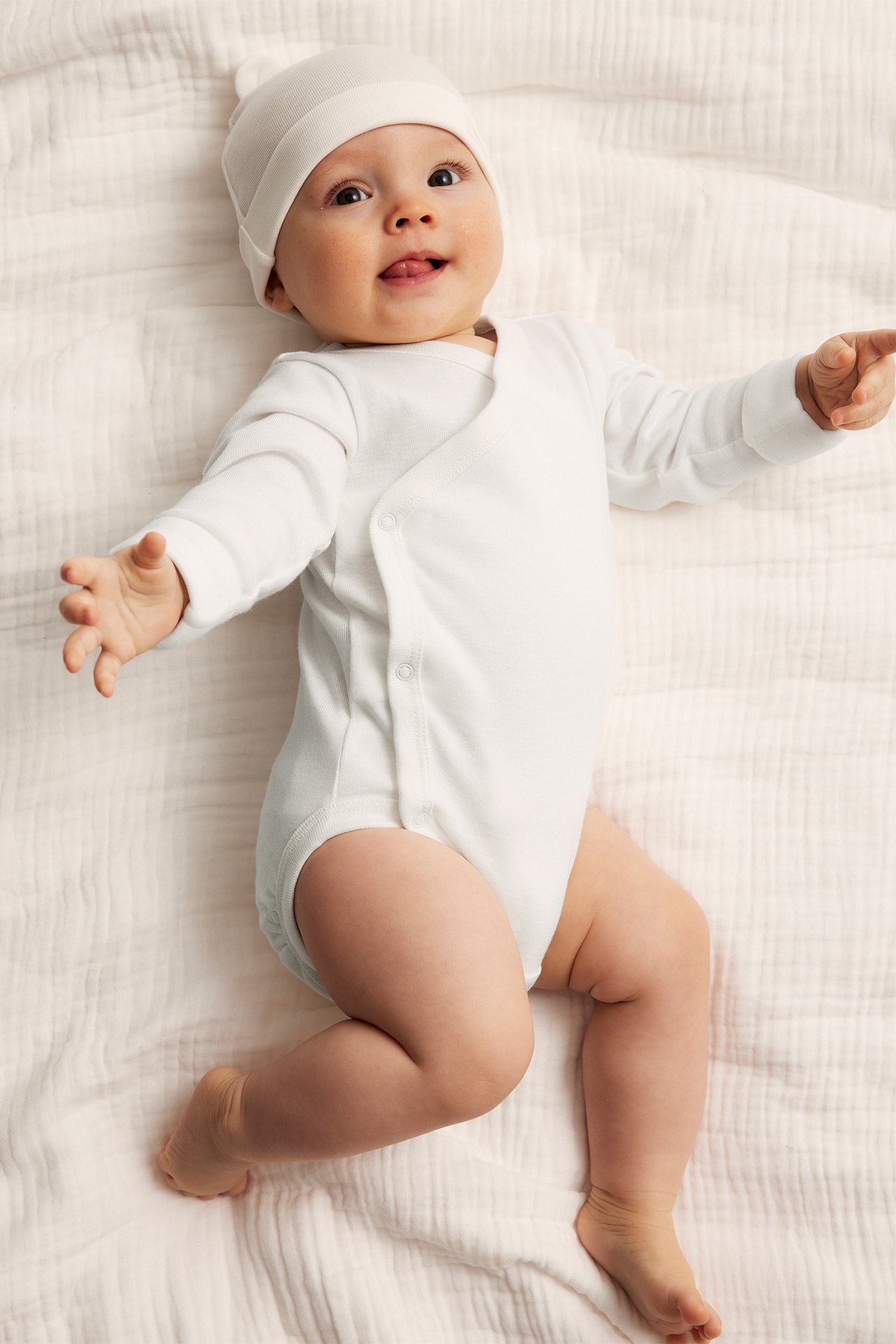 Frontbilde av en baby i en offwhite langermet bomulls-omslagsbody med trykknapper og en matchende lue.