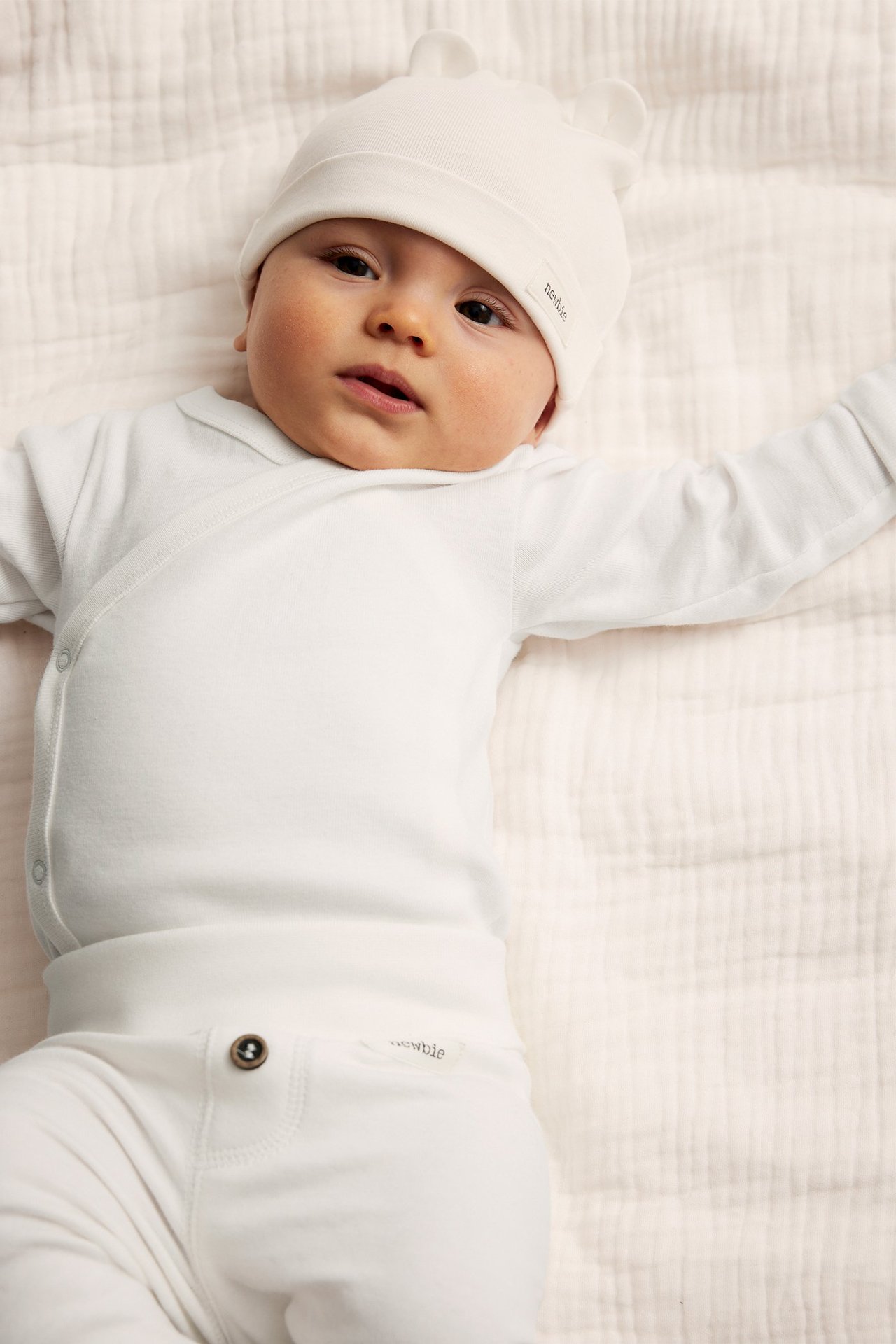 Ovanifrån: Baby i offwhite omlottbody i bomull, matchande mössa med öron och byxor.