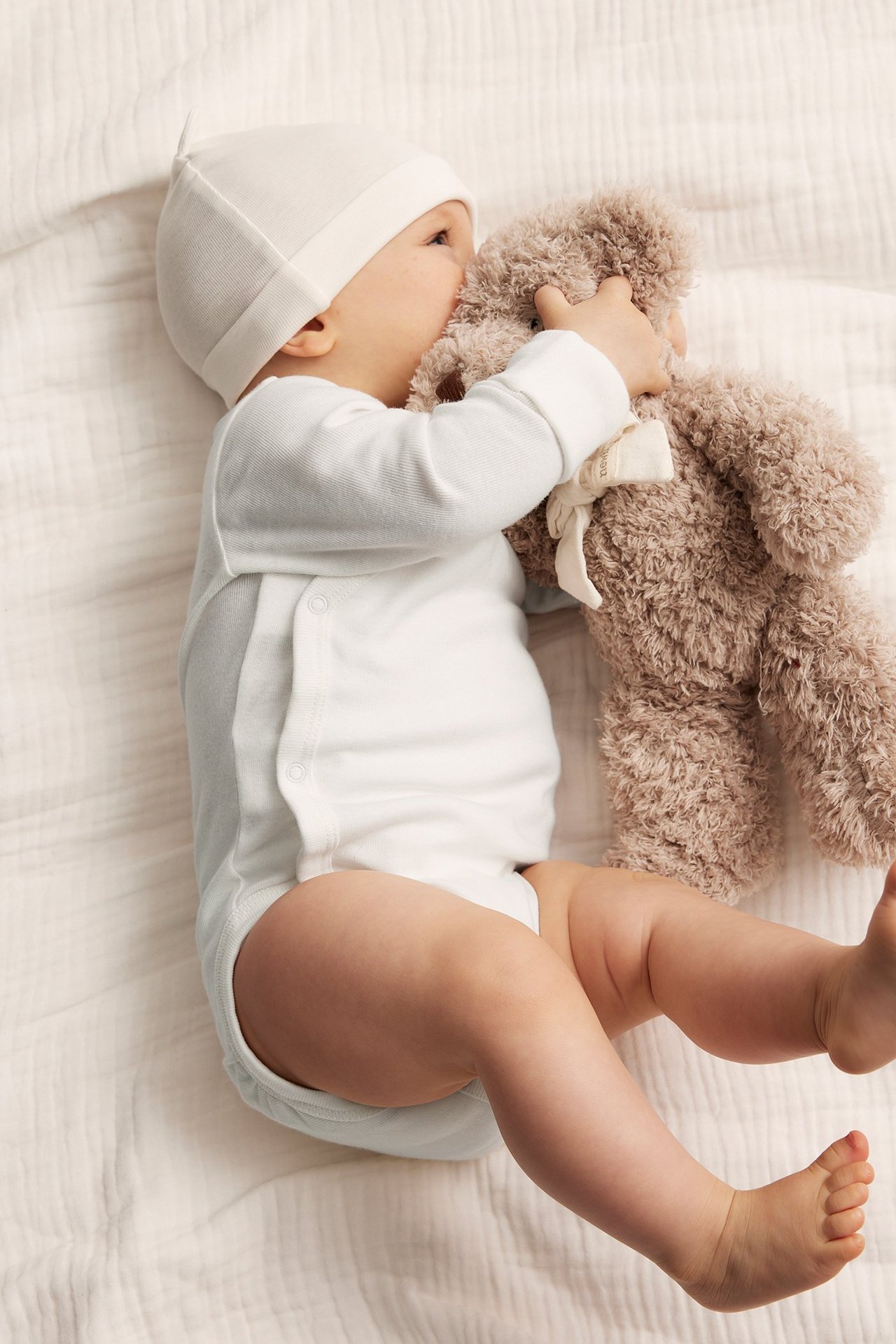 Ovanifrån: Baby i offwhite omlottbody med sidotryckknappar och matchande mössa, håller en nallebjörn.