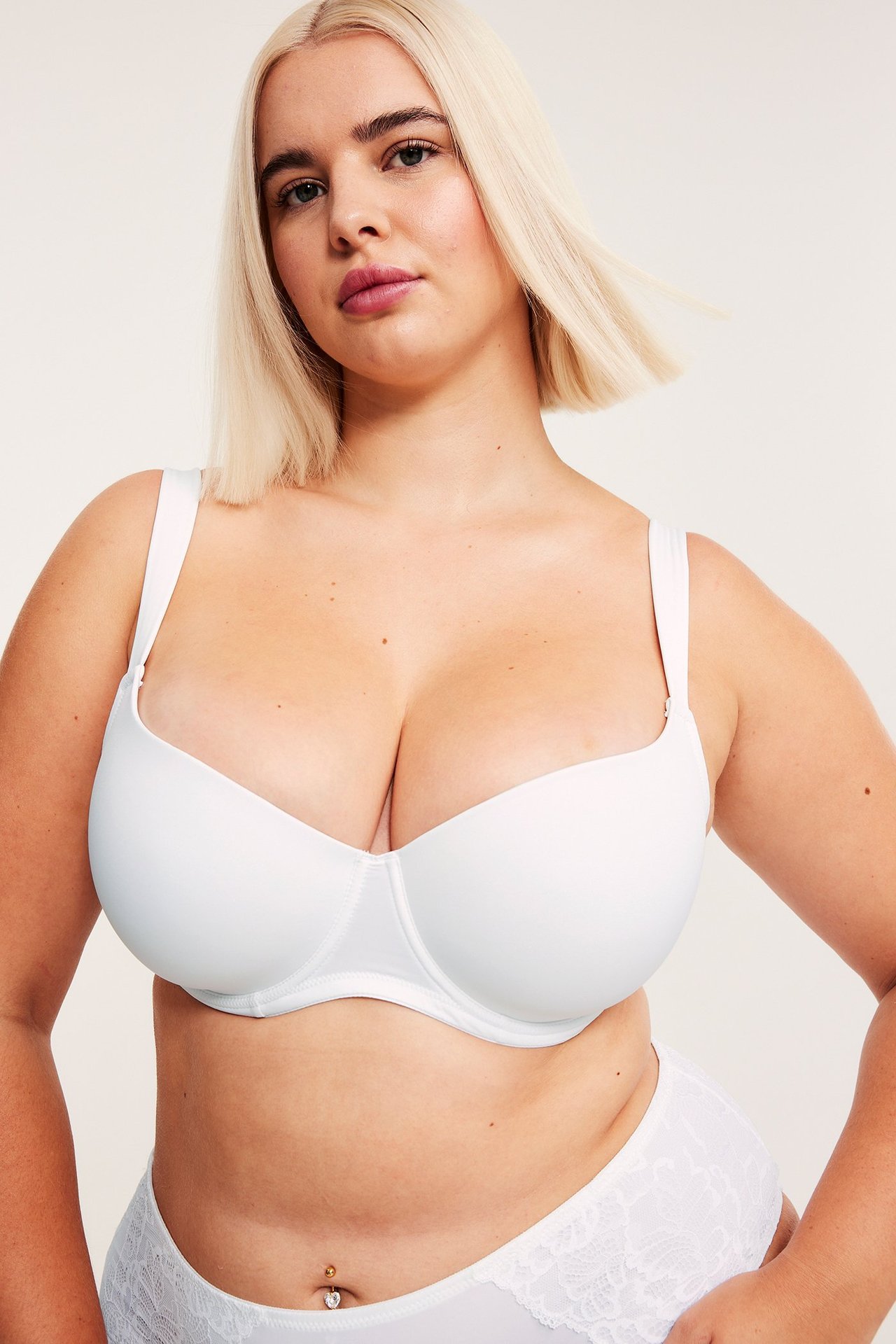 Nainen edestäpäin, yllään valkoiset topatut T-paita-rintaliivit pehmustetuilla olkaimilla ja pitsialushousut.