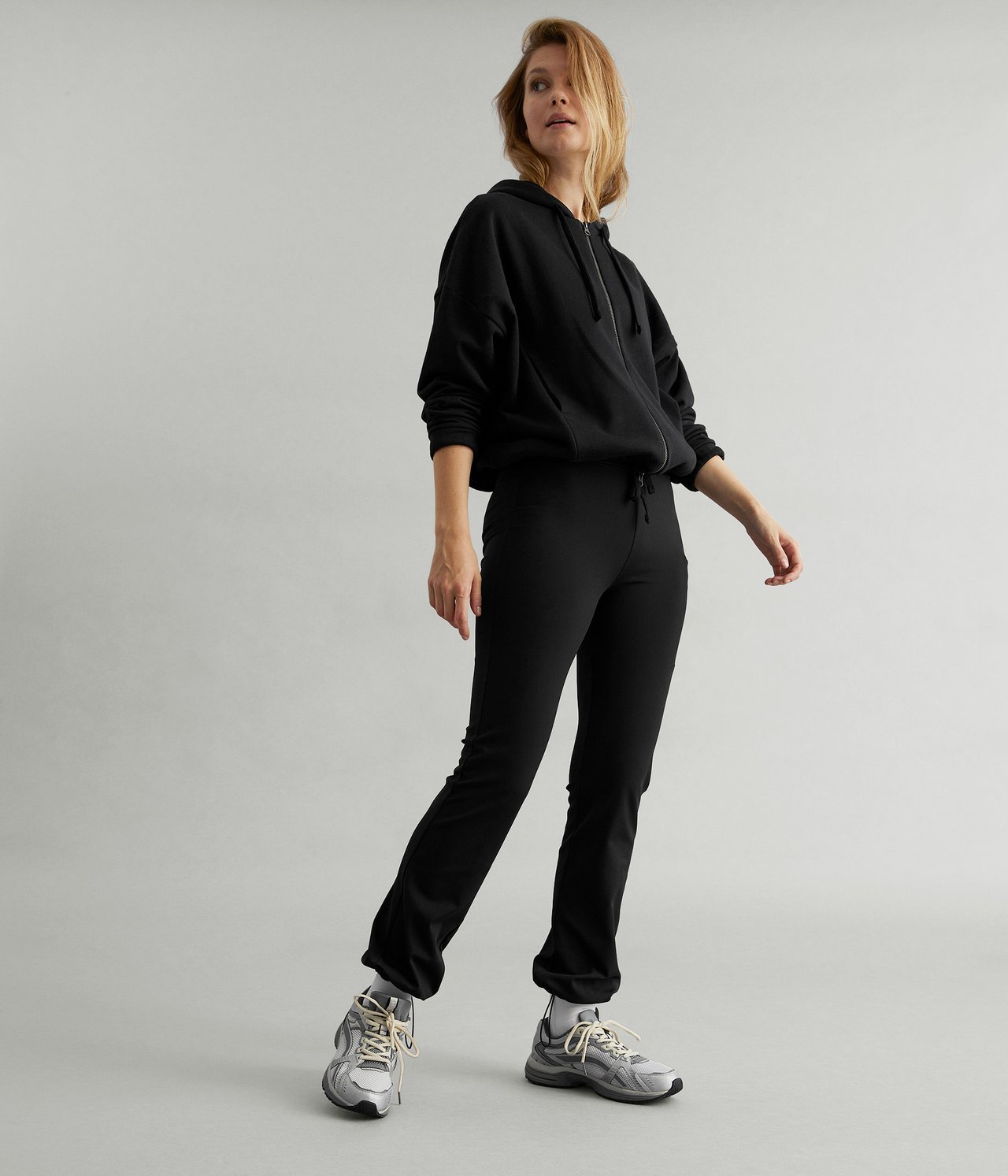 Edestä kuvattu nainen mustissa bootcut-treenitrikoissa, mustassa hupparissa ja harmaissa lenkkareissa.