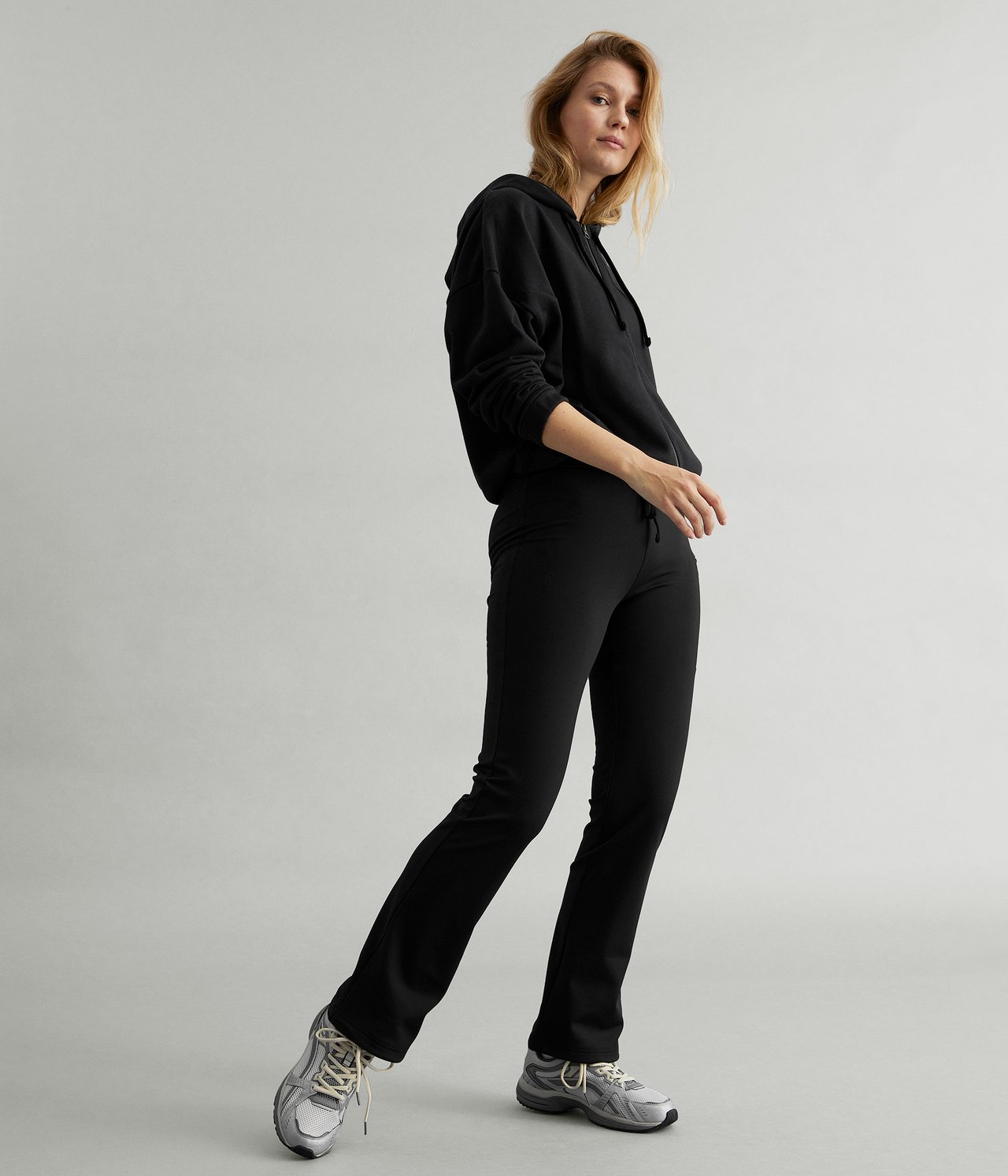 Edestä kuvattu nainen mustissa bootcut-treenitrikoissa, joissa kiristysnyöri helmassa, mustassa hupparissa ja harmaissa lenkkareissa.