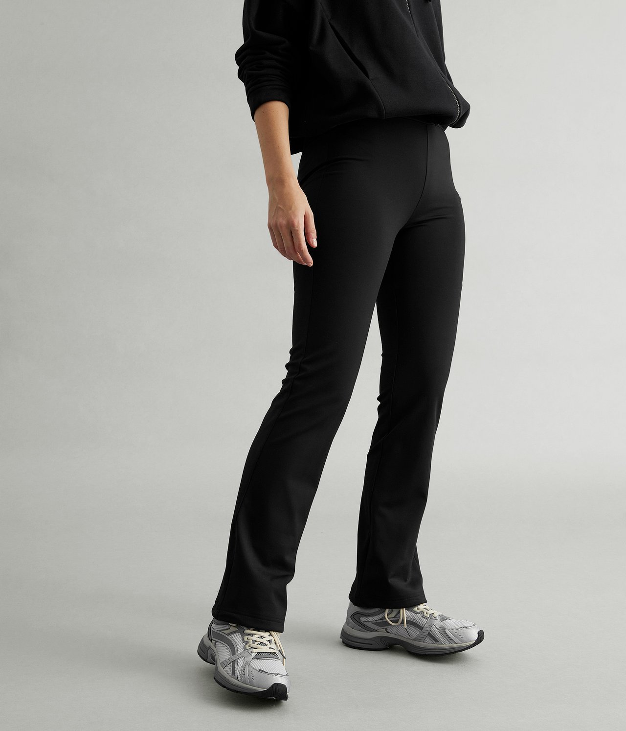 Edestäpäin: nainen mustissa korkeavyötäröisissä bootcut-treenitrikoissa, mustassa topissa ja harmaissa urheilukengissä.