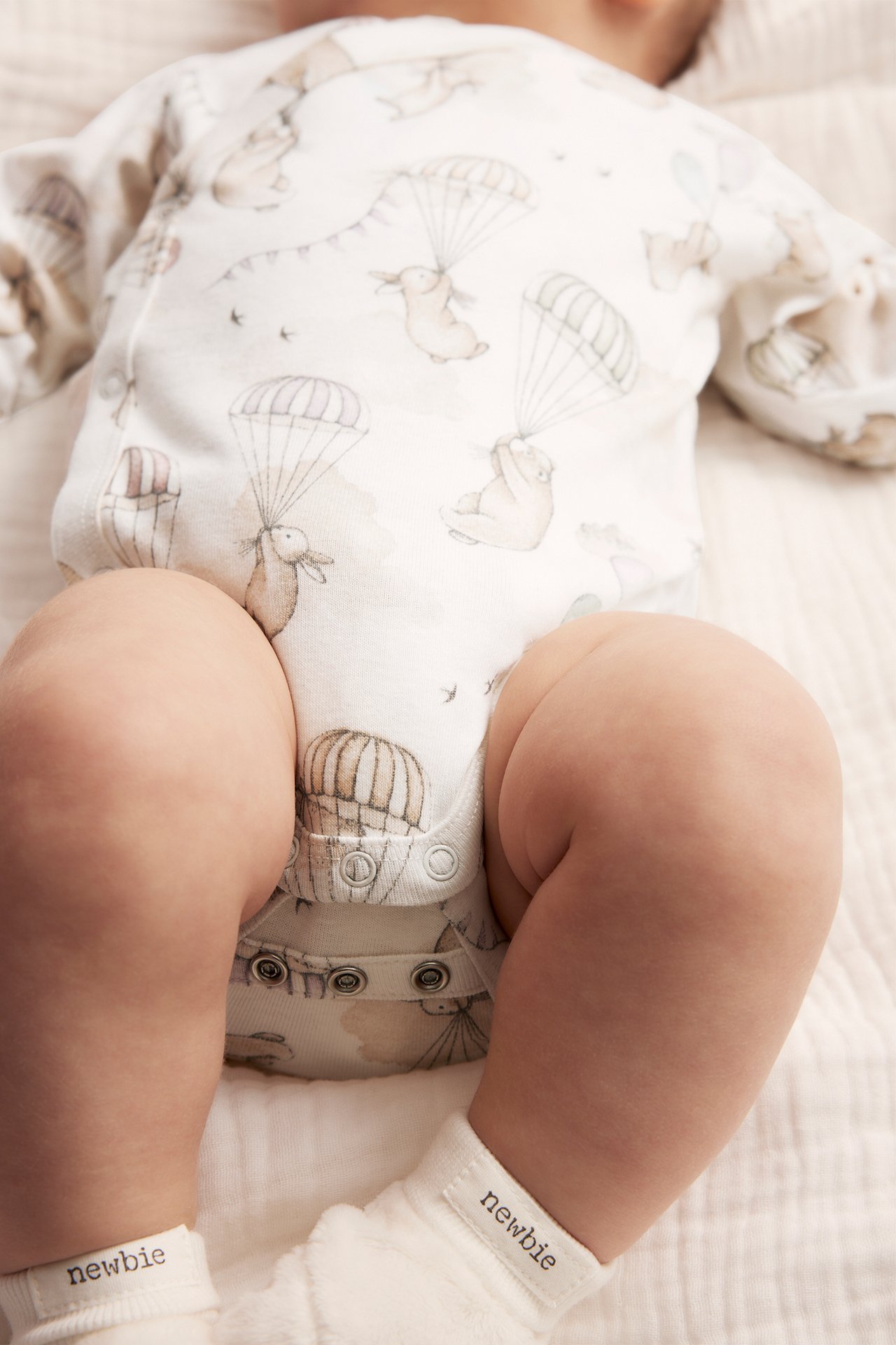 Ylhäältä kuvattu vauva luonnonvalkoisessa kuviollisessa kietaisubodyssä laskuvarjoeläimillä, valkoiset sukat.