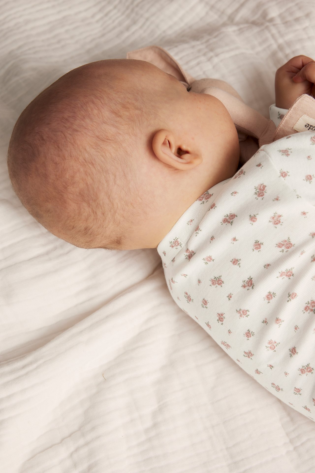 Lähikuva, sivukuva vauvasta yllään luonnonvalkoinen kukkakuvioinen pitkähihainen kietaisubody, pitäen vaaleanpunaista kangasta.