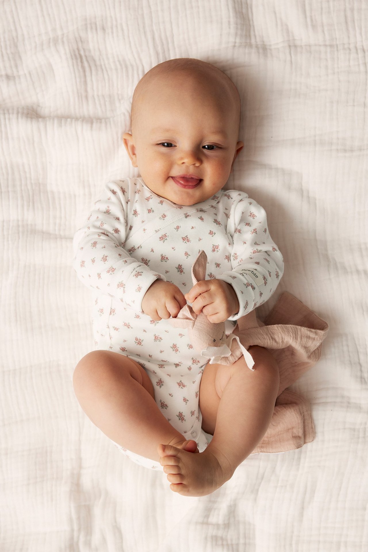 Uppifrån-vy av leende baby i offwhite blommig omlottbody med lång ärm, håller rosa kanin-snuttefilt.