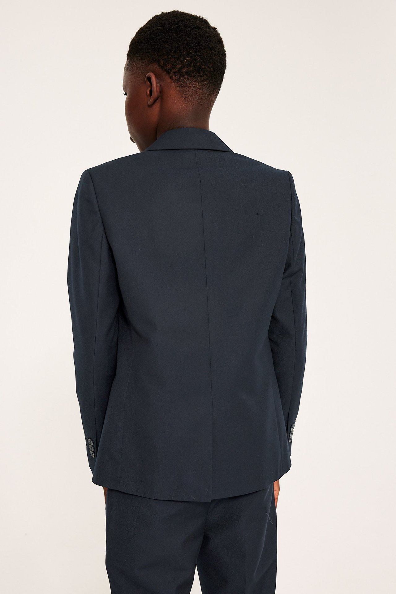 Ryggvisning av et barn i mørkeblå blazer med mansjettknapper, og matchende bukser.