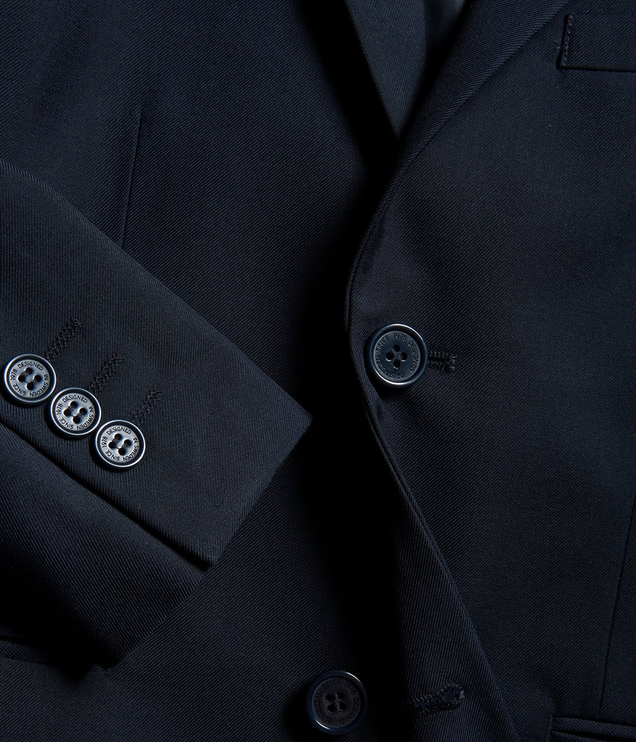Zbliżenie na granatową, jednolitą marynarkę z zapięciem na guziki z przodu i trzema guzikami na mankiecie.