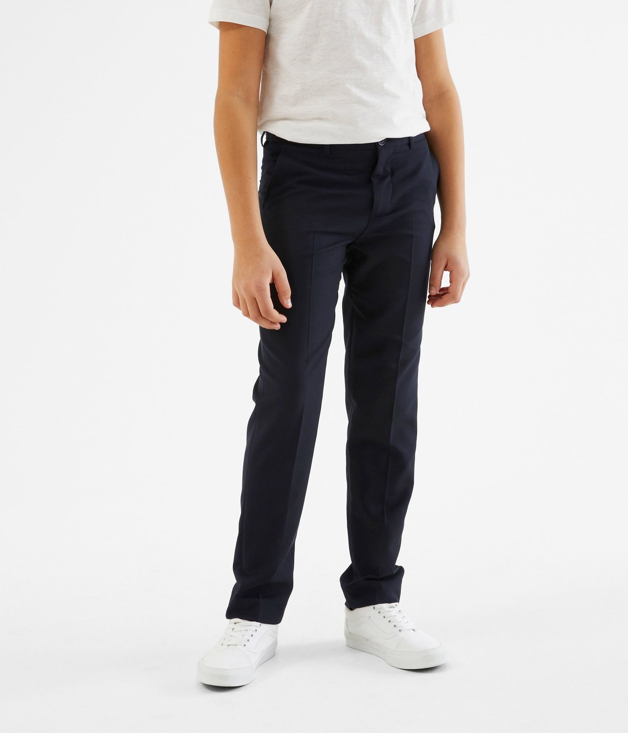 Edestä kuvattu lapsi tummansinisissä puvunhousuissa, valkoisessa t-paidassa ja valkoisissa tennareissa.