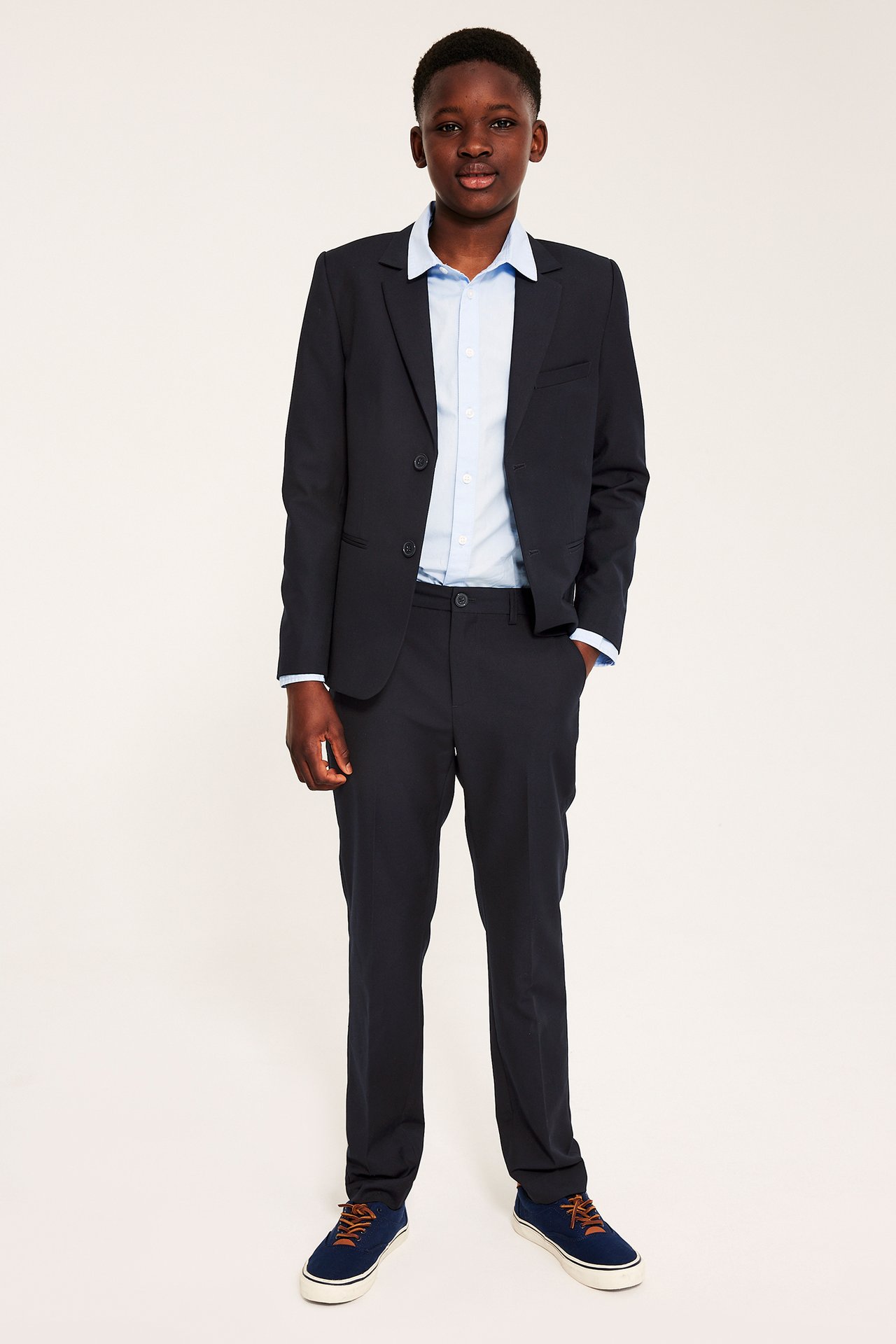 Etunäkymä: lapsi tummansinisissä pukuhousuissa, yhteensopivassa takissa, vaaleansinisessä paidassa ja sinisissä lenkkareissa.