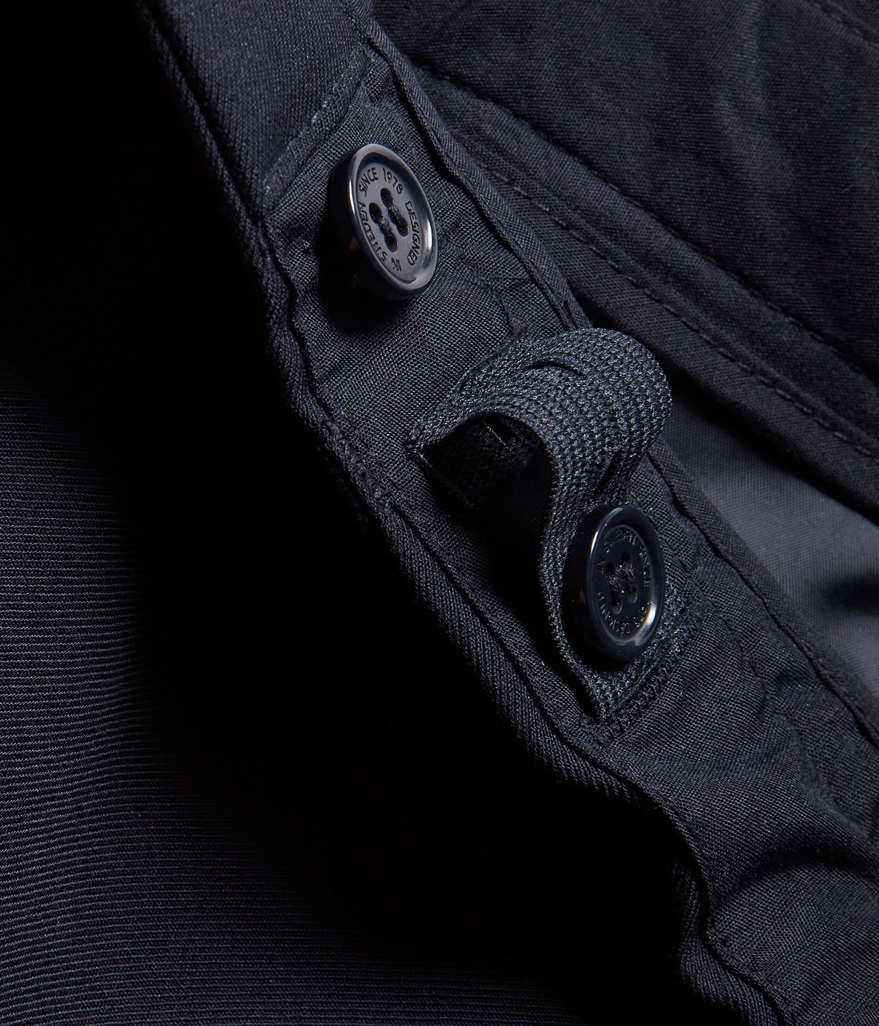 Zbliżenie na ciemnoniebieskie spodnie garniturowe, ukazujące srebrny guzik z napisem i materiałową szlufkę.