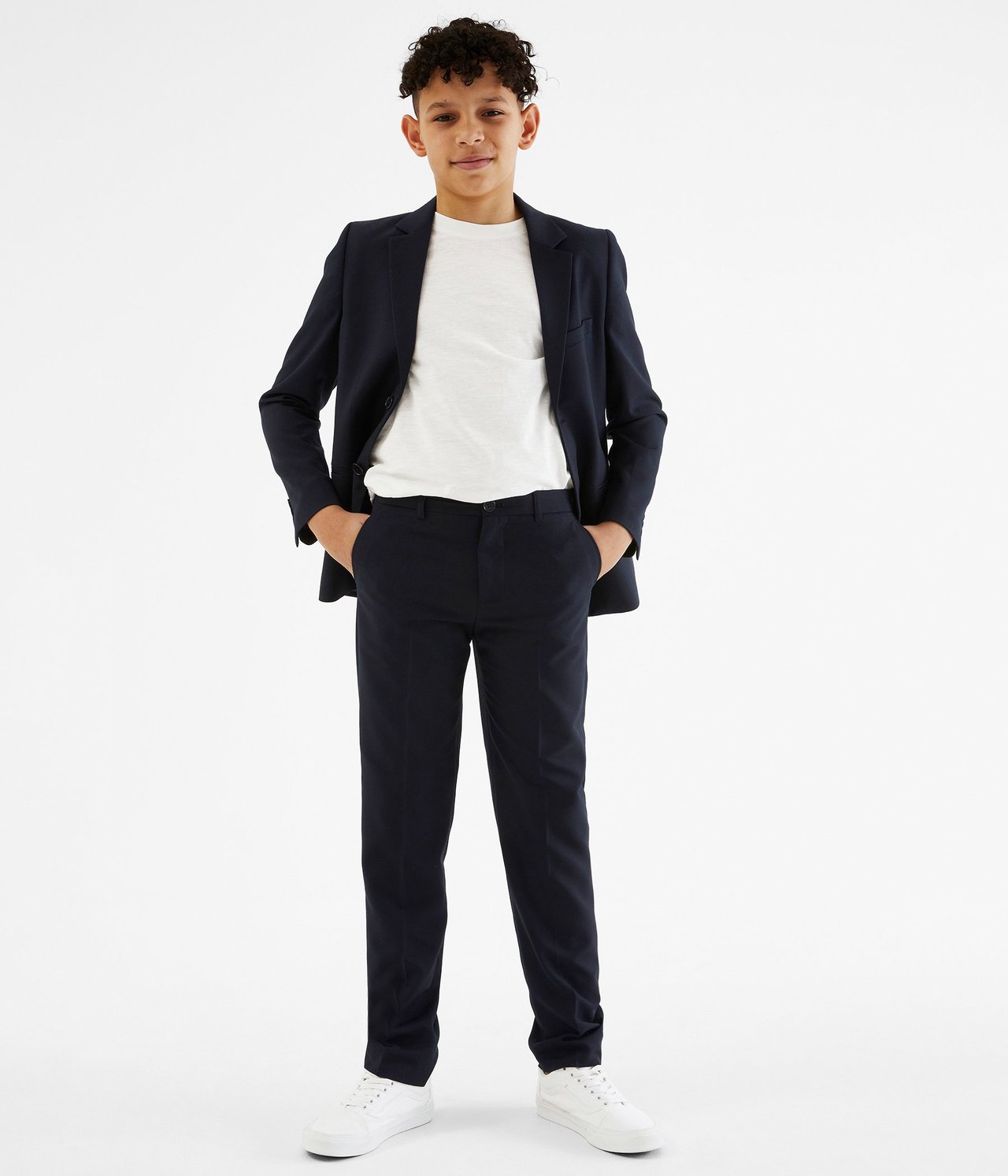 Edestä, lapsi tummansinisissä puvunhousuissa, pikkutakissa, valkoisessa t-paidassa ja valkoisissa lenkkareissa.