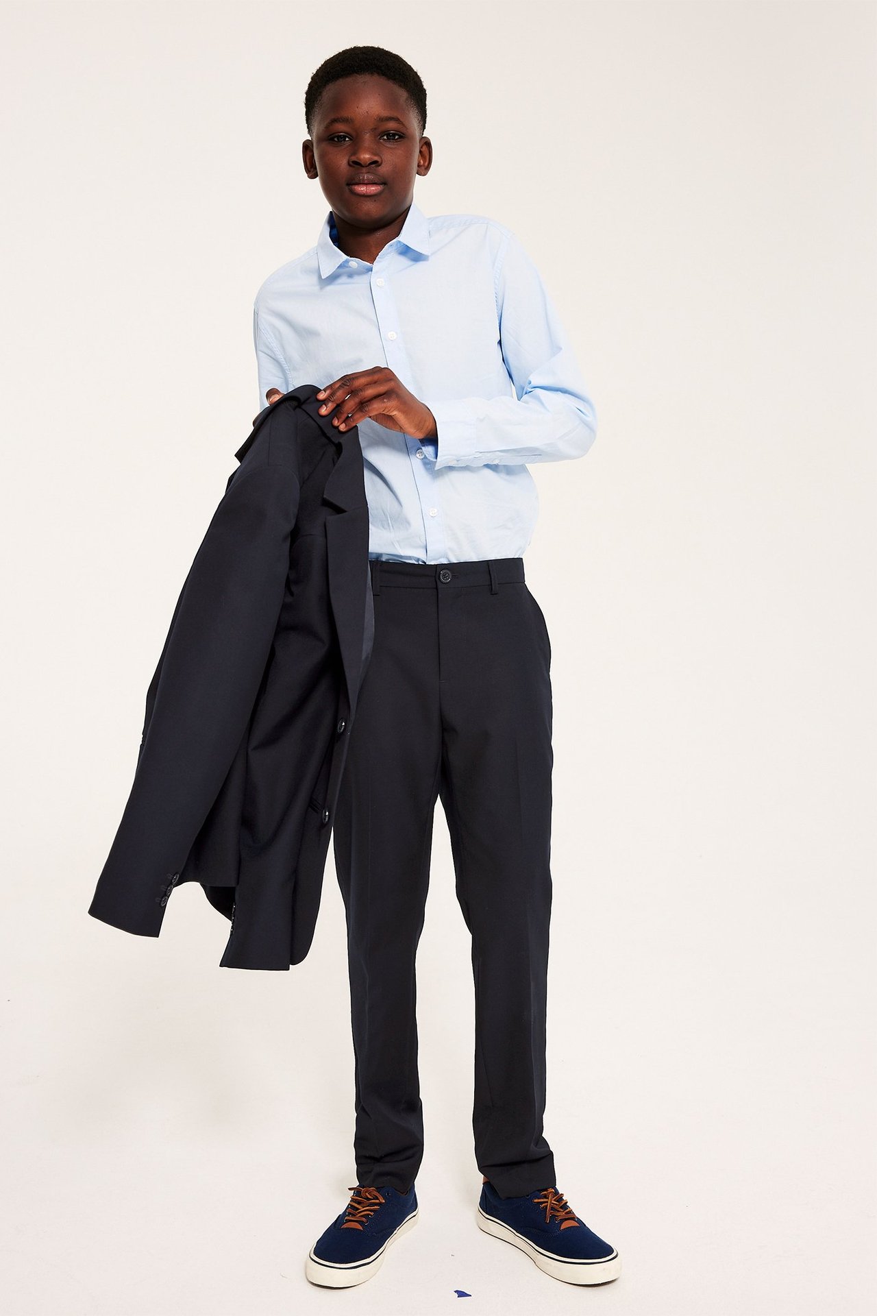 Chłopiec w ciemnoniebieskich spodniach, jasnoniebieskiej koszuli, ciemnoniebieskich trampkach, trzyma ciemnoniebieską marynarkę.